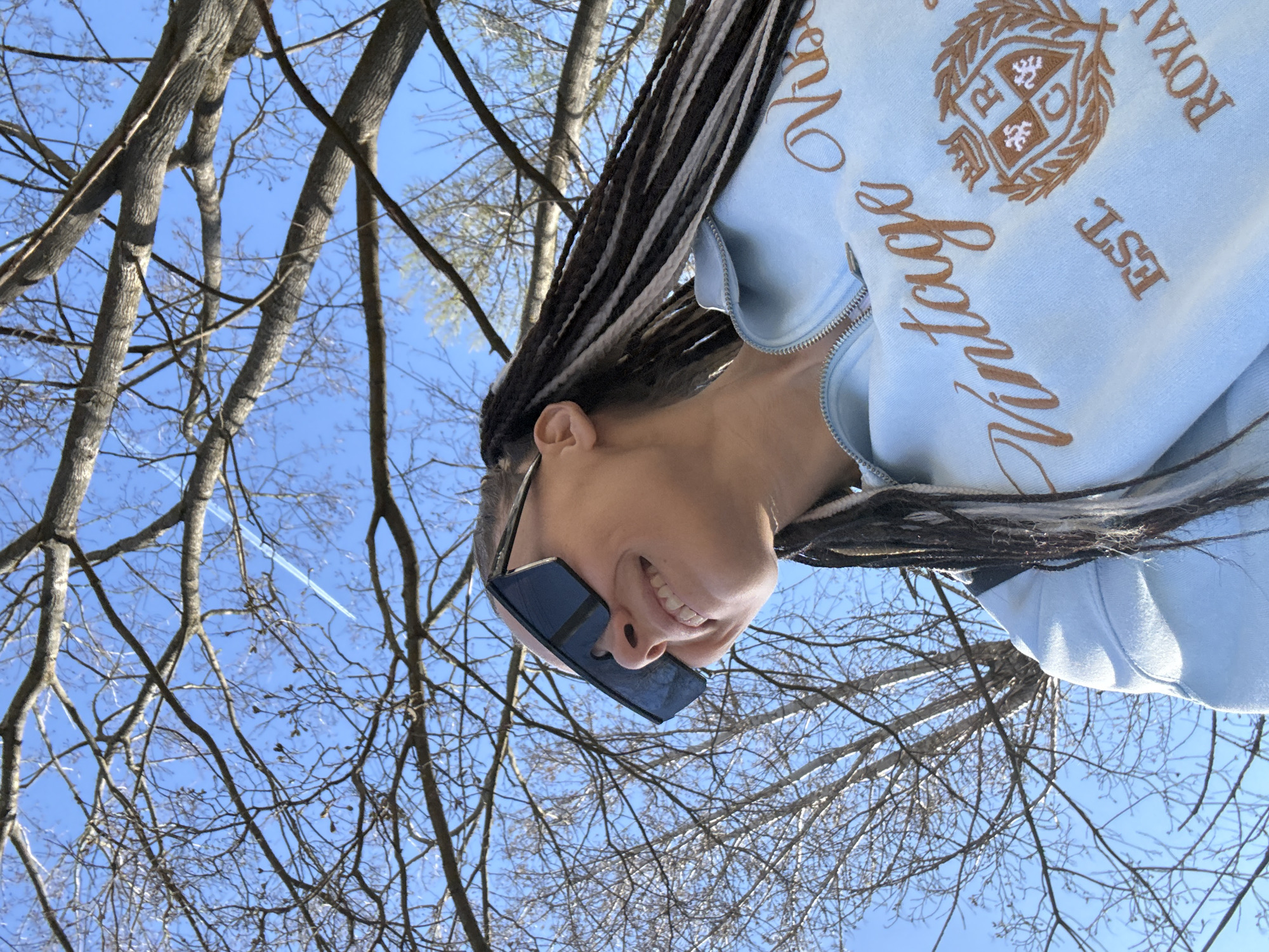 This image, rotated approximately 90 degrees clockwise, captures a cheerful person looking upwards against a backdrop of a bright blue sky and bare tree branches. The person, positioned in the foreground, has a medium skin tone and is smiling broadly, revealing their teeth. They are wearing rectangular, dark-lensed sunglasses. Their hair is styled in long, distinct braids with a striking pattern of alternating dark brown and lighter (possibly white or light grey) sections.
The individual is wearing a light blue zippered jacket or hoodie. Prominently embroidered on the garment in brown thread are the cursive word "Vintage," partial words like "ROYAL" and "EST.," and an ornate crest featuring what appear to be lion-like figures and the letters "R" and "Q."
The background reveals a clear, vibrant blue sky, indicating bright, sunny daytime conditions. A dense network of bare, leafless deciduous tree branches crisscrosses the sky, suggesting the season is late winter or early spring. A faint white contrail is visible streaking across the sky in the upper left background (when viewed upright). The overall mood of the image is joyful and relaxed, set outdoors in what appears to be Rusanovo, Russia.