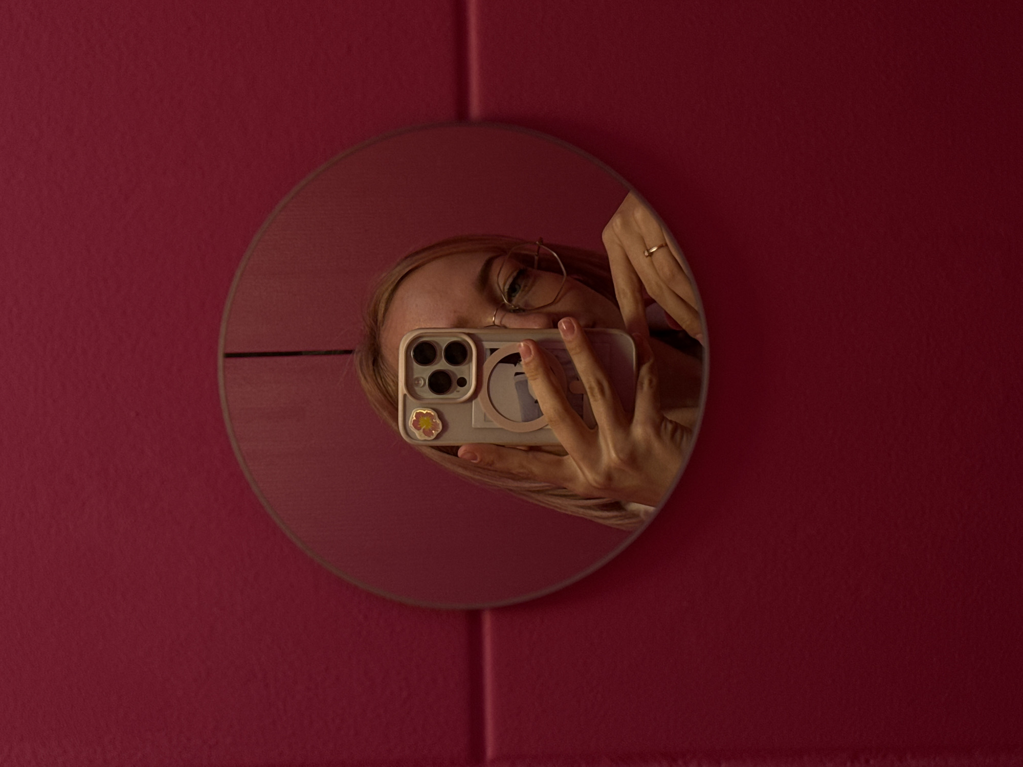 The image shows a selfie taken in a round mirror hanging on a pink wall. The mirror reflects a person holding a smartphone with a camera visible. The phone has stickers on the back. The person is wearing glasses and a ring. The wall appears to have vertical seams, possibly indicating it is made of panels. The location is not discernible from the image.