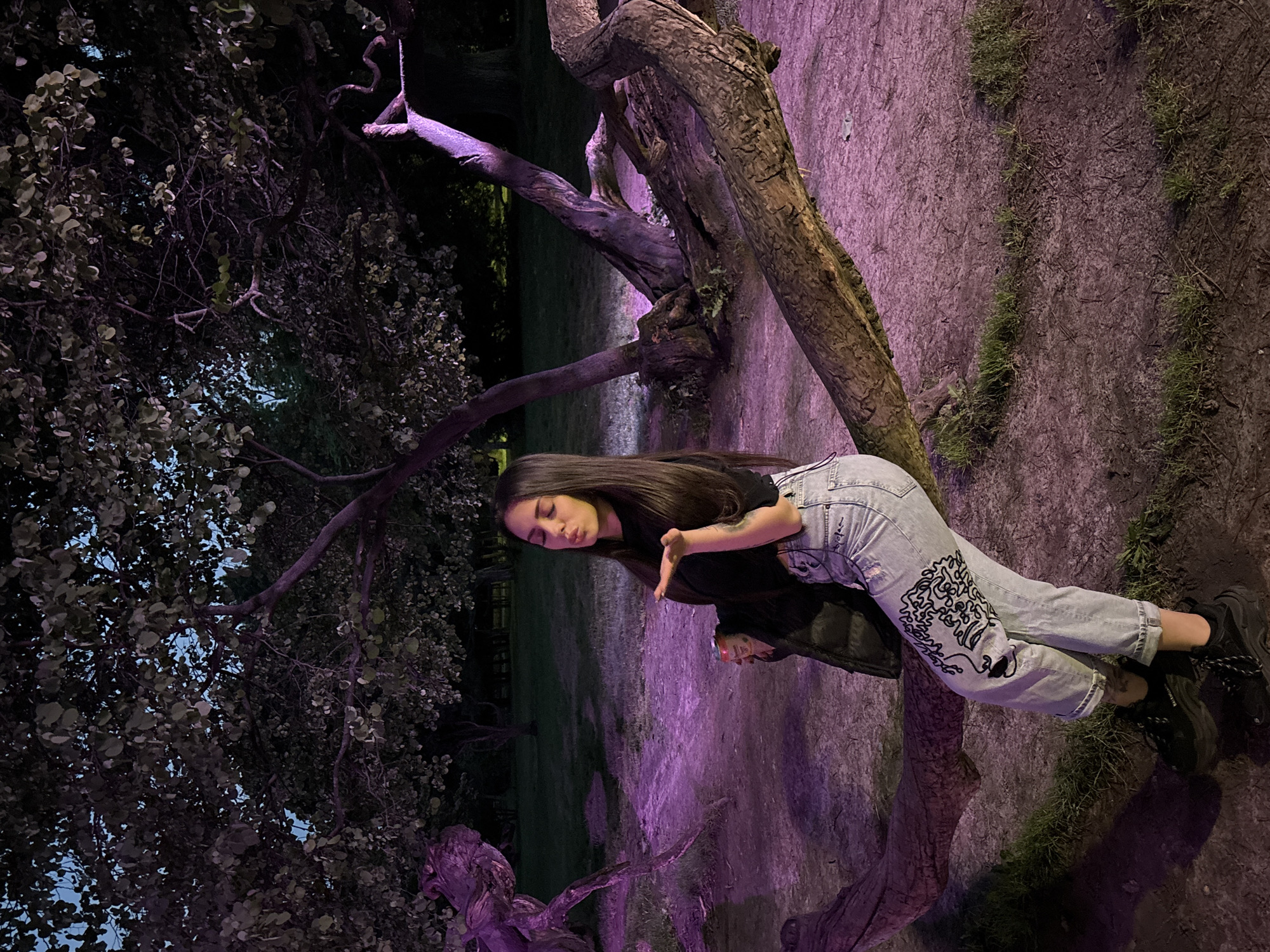 This image captures a young woman posing in a dimly lit outdoor environment, likely a park, at night in Varna, Bulgaria. She has long, dark hair and is dressed in a black t-shirt, light blue ripped jeans adorned with distinctive black swirling patterns on the left leg, and black sneakers. She leans casually against a thick, gnarled tree trunk, with her right hand raised in a gestural pose and her lips slightly pursed as if blowing a kiss or pouting.
The scene is enveloped in artificial purple or pink light, which brightly illuminates parts of the tree trunks, branches, and the earthy ground. This creates a striking contrast with the darker areas of the image, where other trees and foliage appear shadowy against a dark sky. Patches of moss or grass are visible on the ground, also catching the purple light.
Notable details include the intricate black design on her jeans and the brand name "BALENCIAGA" clearly visible on the side of her black sneakers. The dramatic lighting and the model's pose contribute to a moody and artistic ambiance.