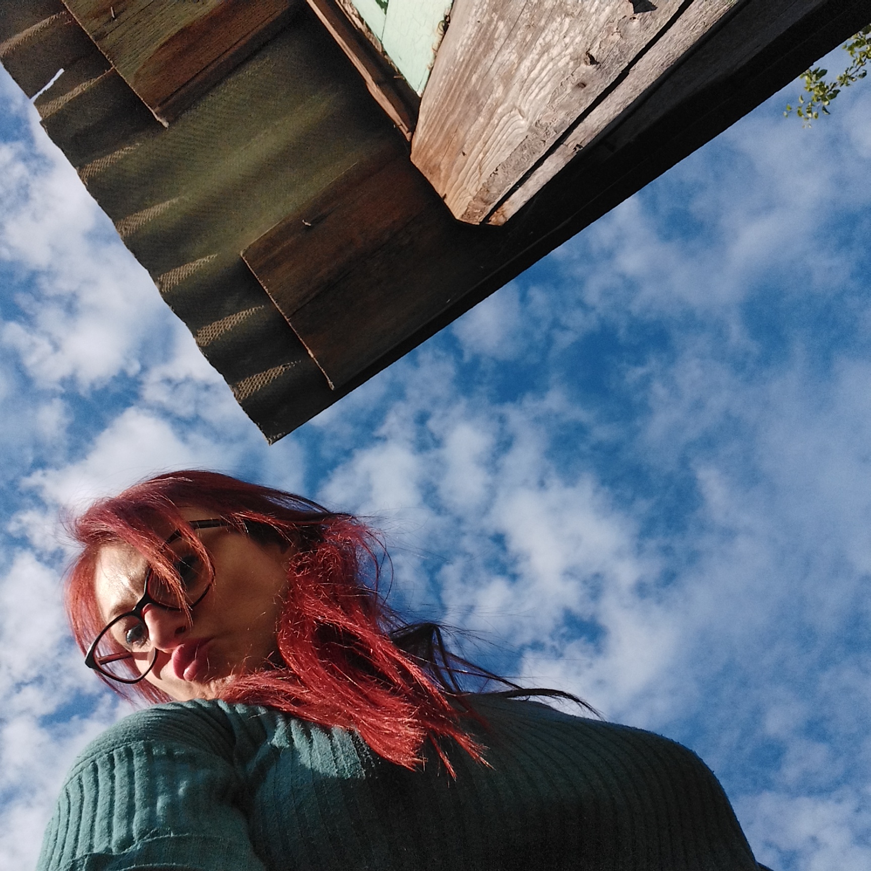 A low-angle, full-body outdoor photograph shows a person with vibrant red hair and glasses looking up at the camera, with a corrugated roof structure above and a blue sky with scattered clouds in the background. The person is wearing a teal sweater with a ribbed texture. The roof appears to be made of weathered wood and corrugated metal. The lighting suggests it is daytime. The scene is outdoors, with no indication of a specific city or location.