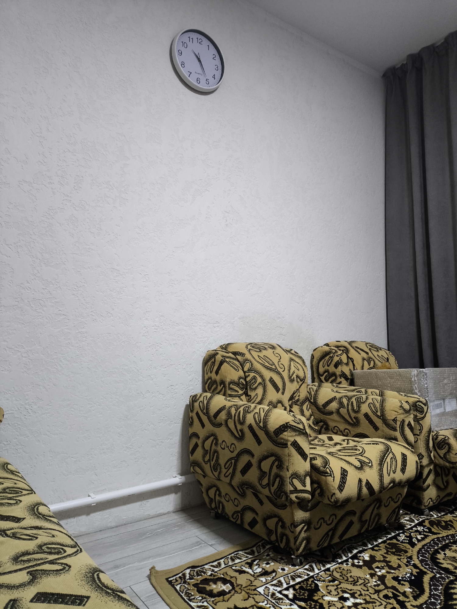 The image depicts an interior scene, likely a living room, in Bălți, Moldova. A light-colored wall with a textured finish occupies the majority of the frame. In the upper left quadrant, a round, white analog clock with black numerals and hands is mounted on the wall, indicating the time is approximately 10:10. To the right, a dark gray curtain hangs from a rod.
In the lower half of the image, a section of a couch or armchair, upholstered in a yellowish fabric with a black abstract pattern, is visible. Another piece of furniture, possibly an extension of the same set, is partially visible on the left. A patterned rug, featuring intricate designs in brown, black, and cream, lies on the floor in front of the furniture. To the right of the armchair, a few stacked boxes wrapped in clear plastic are present. A white pipe runs horizontally along the bottom of the wall, supported by clips. The floor appears to be light-colored laminate or wood. The overall lighting suggests an indoor environment, possibly with artificial light sources. There are no people visible in the image.
