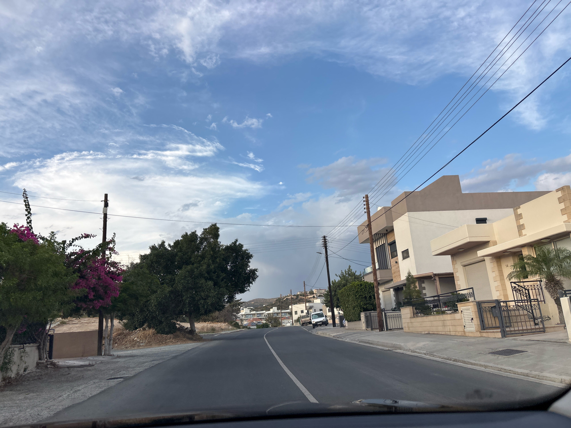 The image captures a street scene in Agios Athanasios Municipality, Cyprus. The road, paved with asphalt and marked with a white dividing line, curves gently to the left. On the right side of the road, modern residential buildings with light-colored facades, geometric designs, and dark metal railings are visible. One building is numbered 56. A palm tree stands in front of one of the houses. Power lines run overhead, supported by wooden and metal poles.
On the left side of the road, lush green trees and vibrant pink bougainvillea add color to the landscape. Further down the street, more trees and buildings are visible, with a hilly terrain in the background. The sky is a mix of blue and white clouds, with a bright, partially cloudy atmosphere. The scene appears to be a residential area on a sunny day. There are no people or animals visible in the image. The view seems to be from inside a vehicle, looking forward.