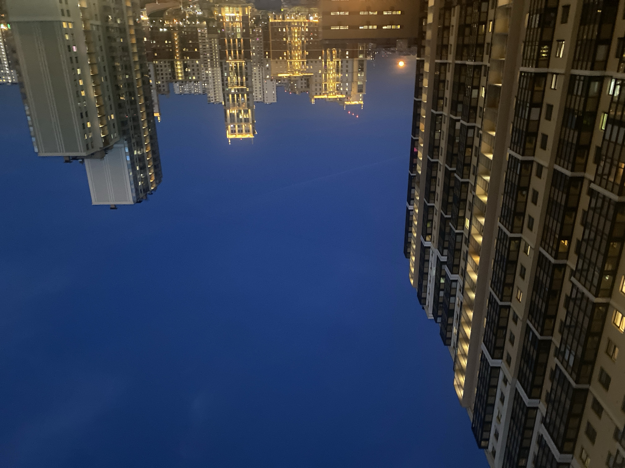 The image captures a vibrant cityscape at night, likely in Saint Petersburg, Russia, characterized by modern high-rise buildings illuminated against a deep blue sky.
In the foreground, a prominent, contemporary high-rise building with a light-colored facade and dark window frames occupies the right side of the frame. Many of its windows emit a warm, yellow light, indicating active residences or offices. Another tall building with similar architectural features is partially visible on the left foreground, also dotted with illuminated windows.
Across the mid-ground and extending into the background, a sprawling urban panorama unfolds. A dense cluster of numerous other multi-story buildings, all brightly lit, creates a rich tapestry of light and shadow, highlighting the extensive nature of the city.
The sky, which dominates a significant portion of the image, is a uniform and deep dark blue, suggesting the time of day is either dusk or late night. A notable detail in the upper-left area of the sky is a distinct, round, yellowish-orange moon, positioned gracefully above the distant urban skyline.
The scene is calm and clear, with no visible clouds. The artificial lights from the buildings are the primary source of illumination, contrasting with the dark sky. No specific human activity is discernible, nor is any visible text present. The overall impression is one of an expansive and bustling city transitioning into the quiet hours of the night.