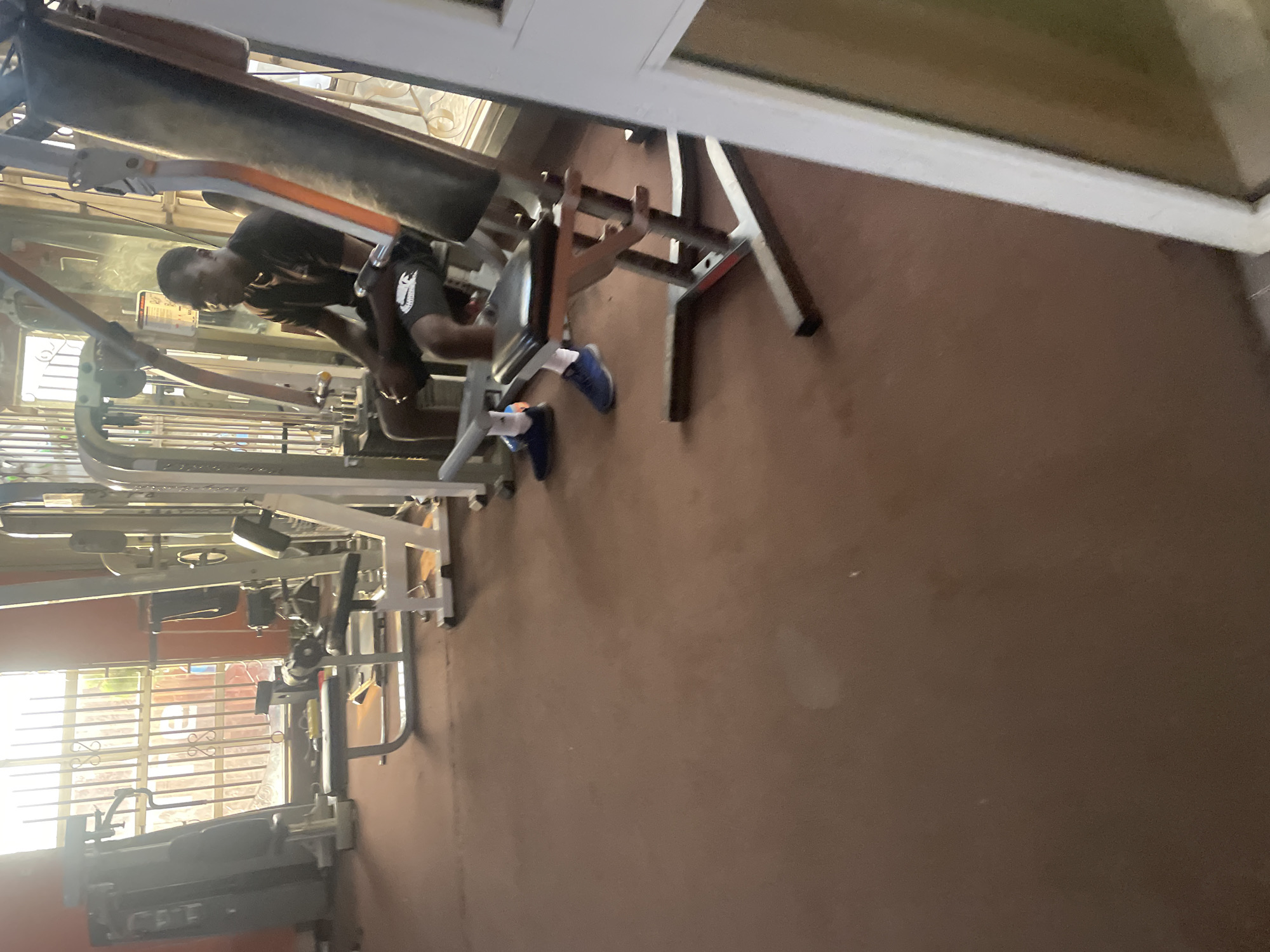 A medium shot captures a young Black man sitting on a weight machine in a gym, likely located in Barakin Akawo, Nigeria. The man is wearing a dark t-shirt and shorts, with blue sneakers and white socks. Sunlight streams through barred windows behind him, illuminating the gym equipment, which includes weight machines and benches. The floor is covered with a reddish-brown mat. The scene appears to be taken during the day, with the sunlight indicating it is likely morning or afternoon. The overall impression is that of a typical gym environment.