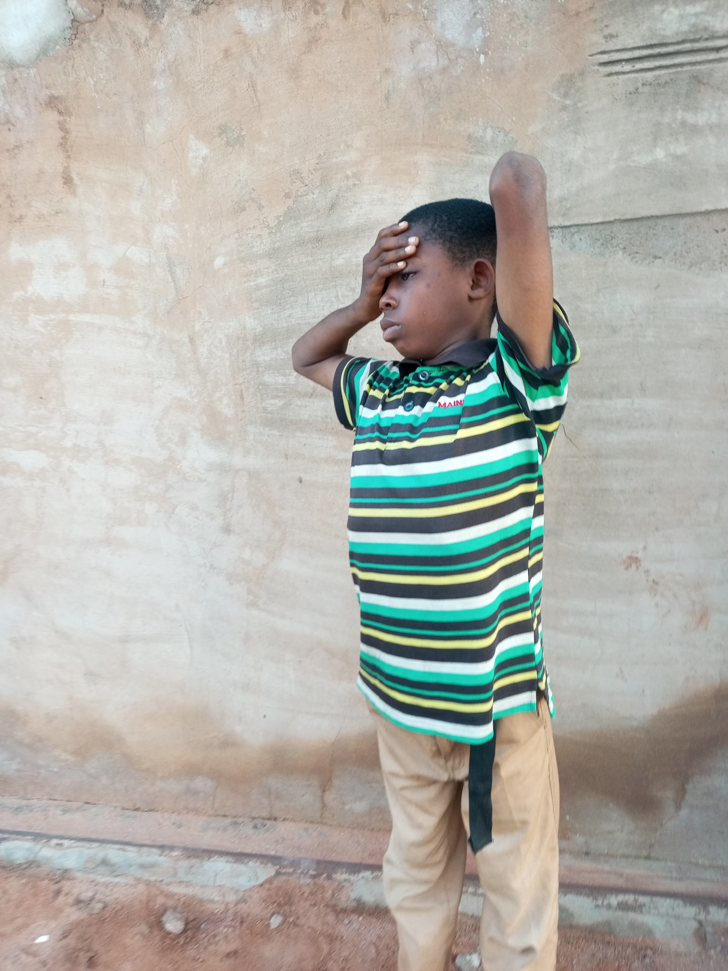 A young boy with dark skin stands against a textured, tan-colored wall. He is wearing a striped polo shirt with horizontal bands of green, white, black, and yellow, and khaki pants. His left arm is bent at the elbow, with his hand covering his forehead, and his right arm is raised behind his head. The boy is looking to his left. The ground in front of him is composed of reddish-brown soil with some small stones. The wall behind him appears weathered, with patches of lighter and darker coloration, and a few horizontal indentations near the top. The scene is outdoors, likely in a residential area in Tempure, Nigeria.