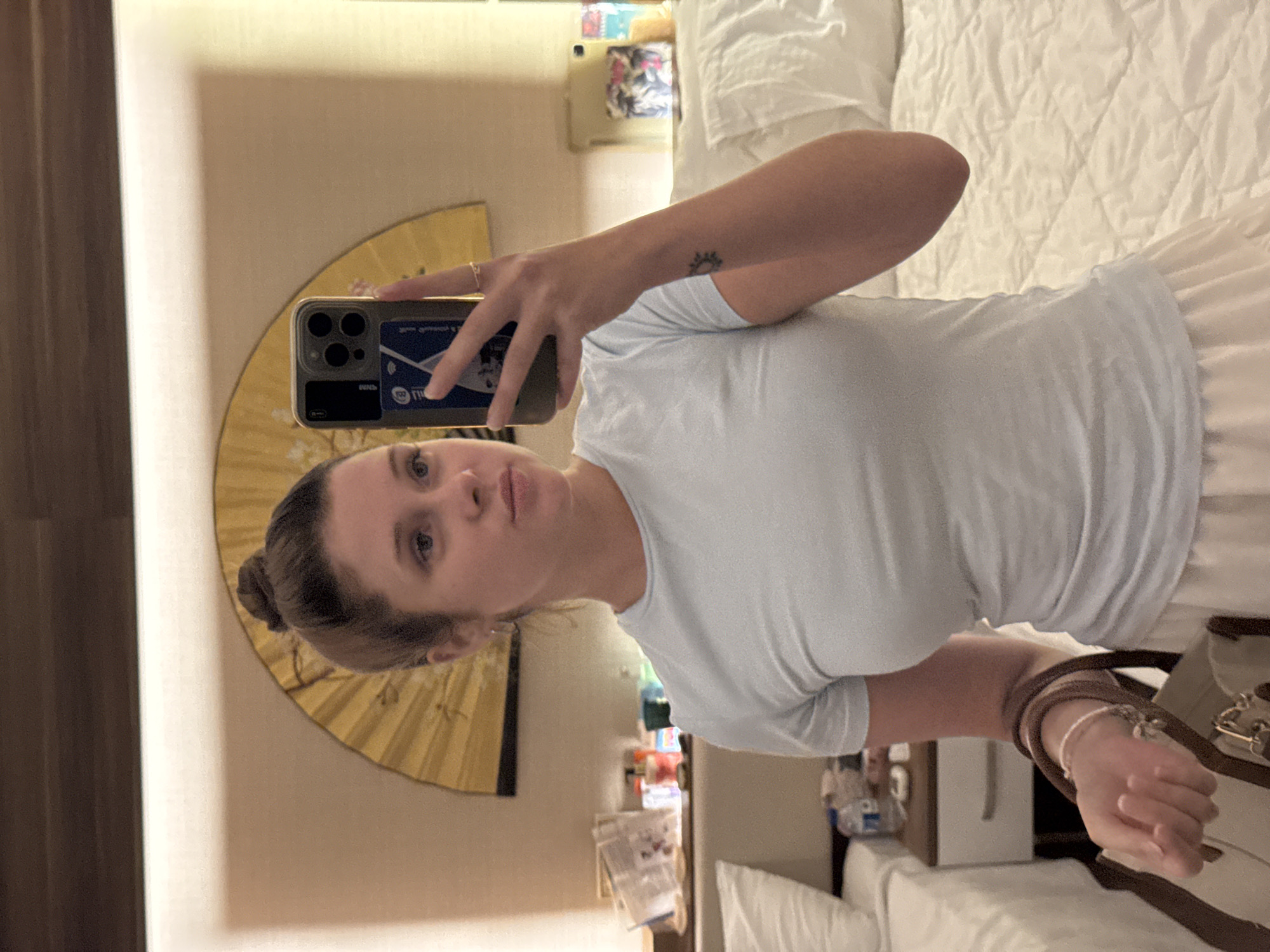 A woman with her hair in a bun, wearing a light blue t-shirt and a white skirt, takes a mirror selfie in a hotel room. She is holding a purse and a smartphone. In the background, there is a bed with white bedding, a nightstand with various items, and a decorative fan on the wall. The scene appears to be indoors, possibly in a hotel. There is no indication of the location being Aksu, Turkey.