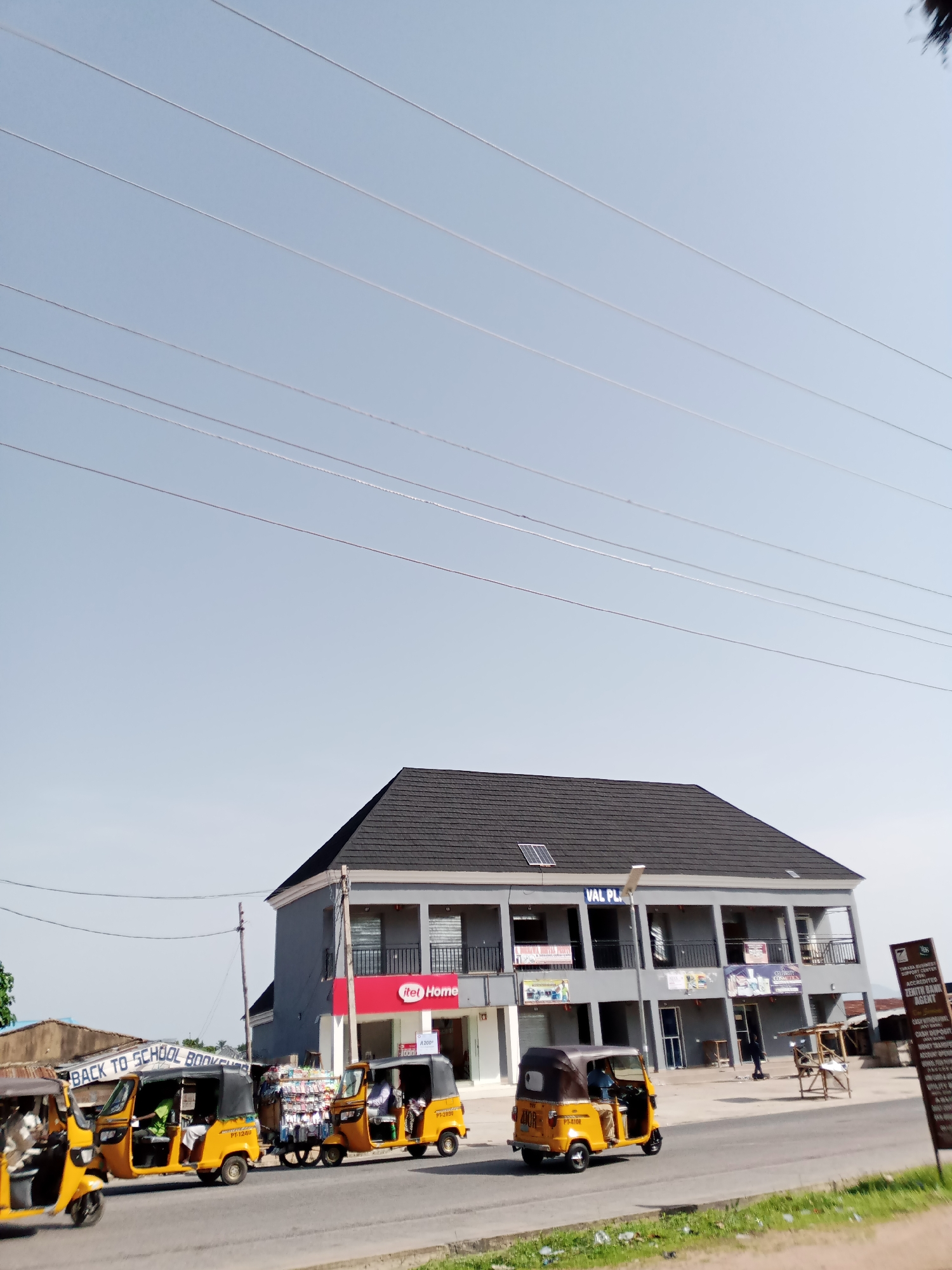 In the city of Jalingo, Nigeria, a street scene unfolds under a clear sky crisscrossed by power lines. A two-story building with a dark, pitched roof stands prominently on the right. Its facade is painted gray, with balconies on the upper level and multiple shop entrances on the ground floor. Signs indicate businesses such as "VAL PLAZA," "itel Home," and "CELEBRITY COSMETICS."
In front of the building, several yellow auto-rickshaws, also known as Keke Napeps, are visible. One is parked near a stall selling various items, while others are in motion on the paved road. A sign reads "BACK TO SCHOOL BOOKSHOP." A young person in a blue shirt is seen driving one of the auto-rickshaws.
To the far right, a tall, dark sign with white text announces "TARABA WOMEN'S COOPERATIVE MOVEMENT SUPPORT CENTER" and "ACCREDITED ZENITH BANK AGENT." Further down the street, another building with a corrugated roof is partially visible. The environment appears to be a commercial area with pedestrian and vehicular traffic.