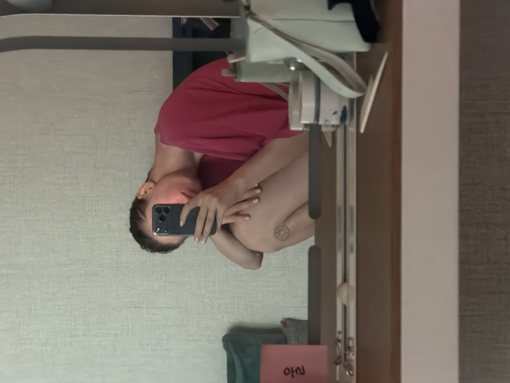 A person is taking a selfie in a mirror, holding a dark-colored smartphone. The person is wearing a bright pink shirt and is seated, with their knees drawn up to their chest. A tattoo of a circular pattern is visible on their left leg. The background appears to be a bedroom with a textured wallpaper. On a wooden surface in front of the mirror, several items are visible, including a pink card with Korean writing, a teal bag, and various containers and objects that suggest a vanity or dressing area. The lighting suggests it is daytime, possibly indoors near a window. The overall scene is casual and personal.