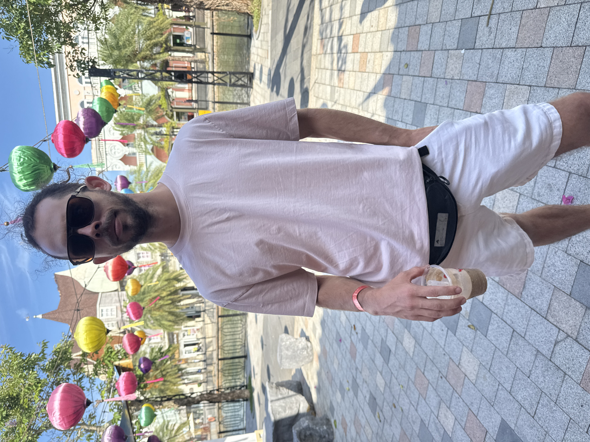 A man with dark hair and sunglasses stands on a paved street, holding a plastic cup with a brown beverage. He wears a light pink t-shirt, white shorts, and a black fanny pack. In the background are colorful hanging lanterns, trees, and buildings with distinctive architecture. The location is in Đà Nẵng, Vietnam. The ground has a pattern of grey, white, and tan tiles. The sky is blue.