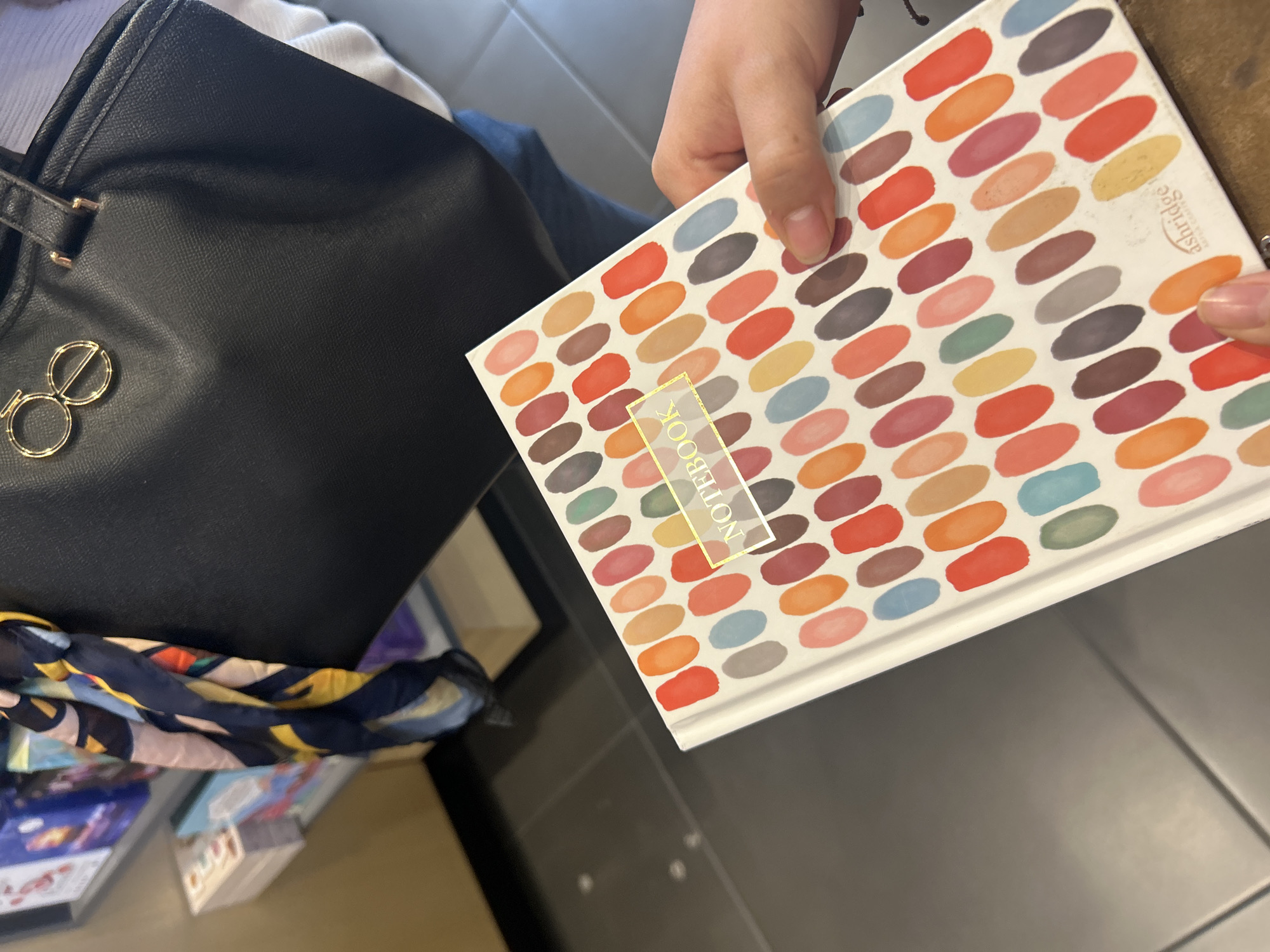 Here is a description of the image:
The image shows a person holding a notebook with an abstract pattern of colorful oval shapes. The notebook has a white background and a gold label that says "Notebook." The person's hand is visible, holding the notebook. Beside the notebook is a black handbag with a gold emblem. A colorful scarf or other textile drapes near the bottom of the bag. The background consists of a gray tiled surface and a glimpse of some packaged items.