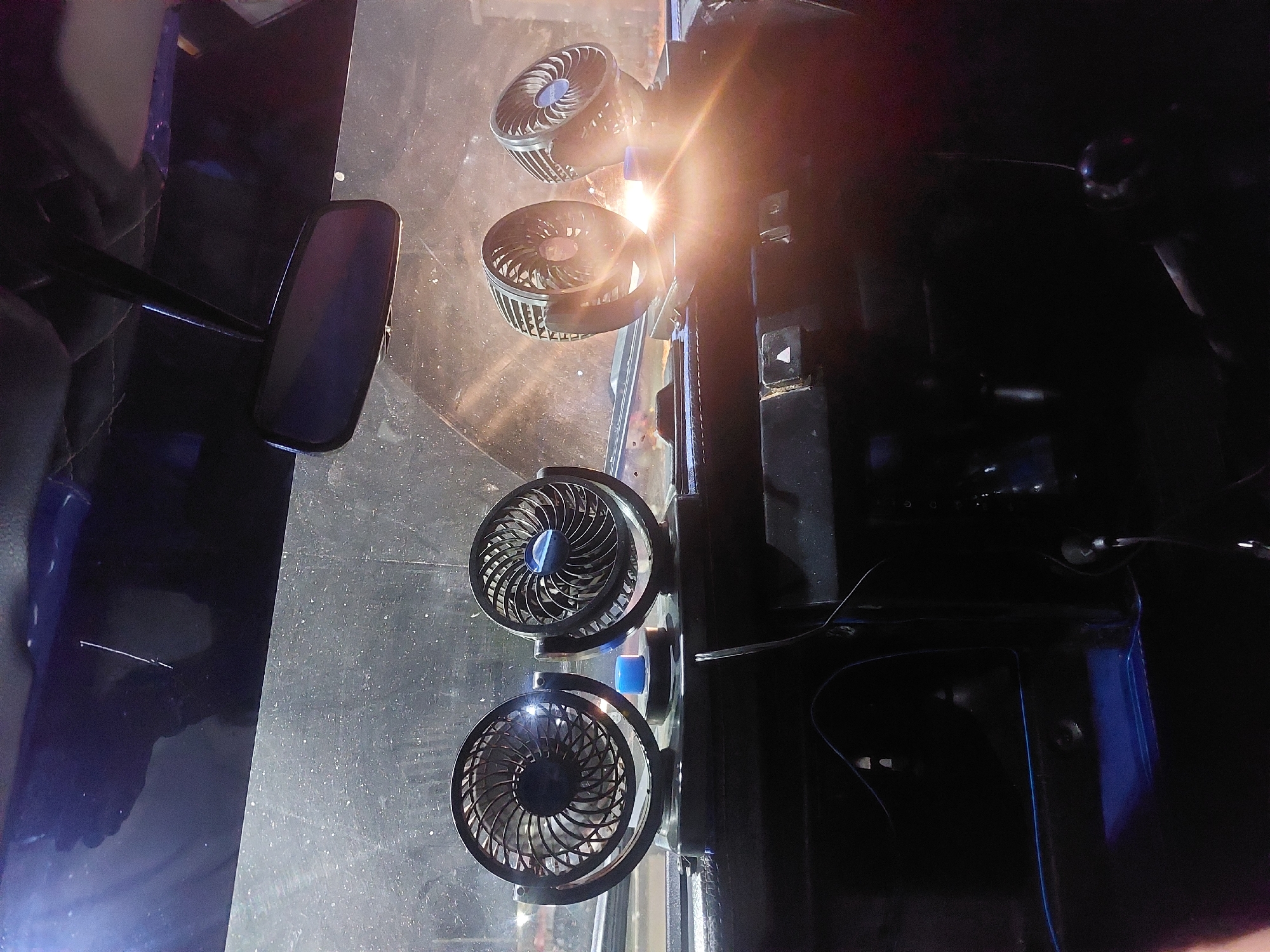 The image shows the interior of a vehicle, possibly a truck, with four small electric fans mounted on the dashboard or windshield. The fans are arranged vertically, with the top two closer together than the bottom two. The vehicle has a rearview mirror on the left side of the frame. The lighting suggests it might be daytime, although the details outside the vehicle are obscured by darkness or shadow. There is no information available to determine if this photo was taken in Dar es-Salaam, Tanzania.
