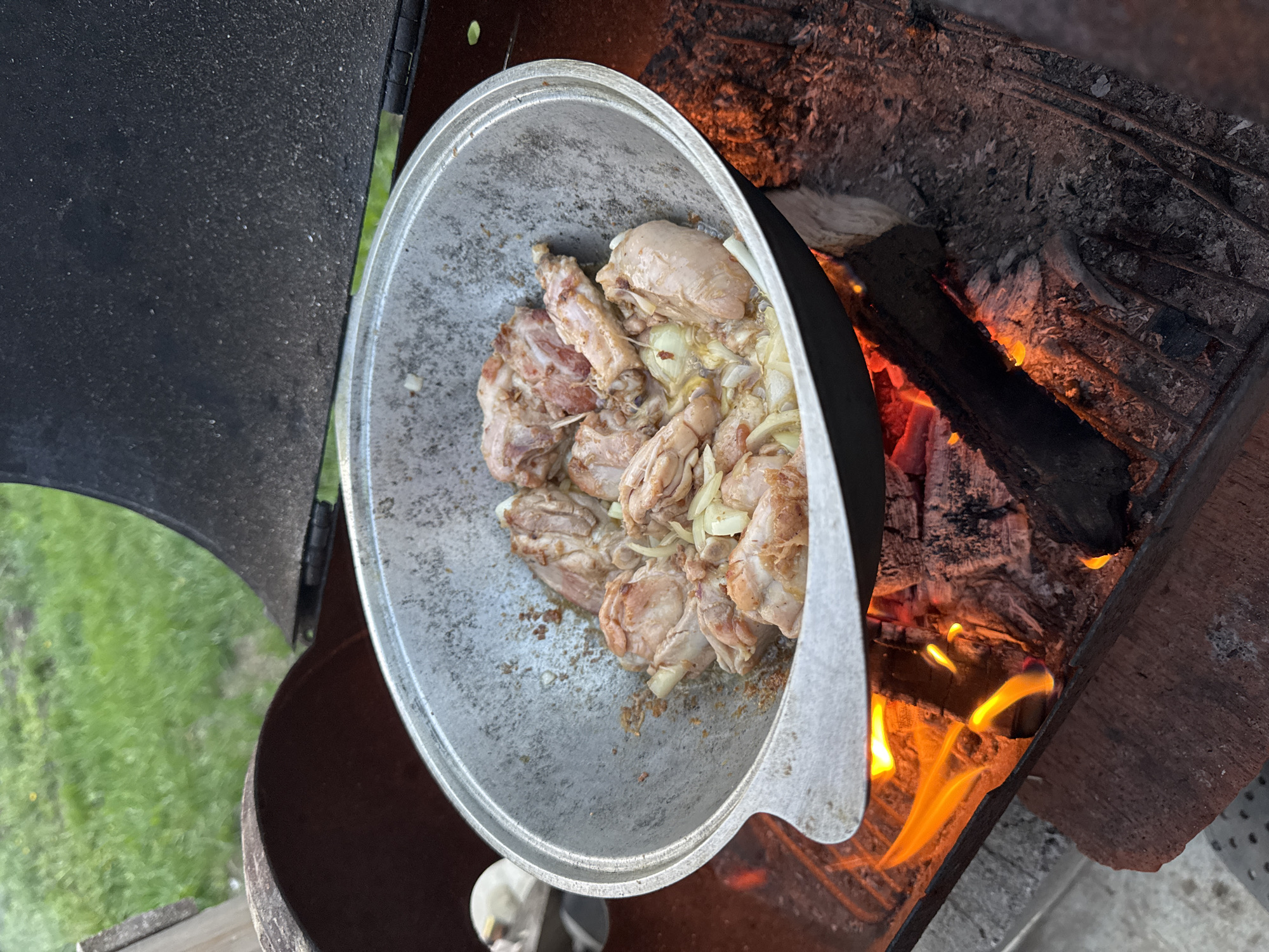 The image captures a cooking scene outdoors. A large, well-worn metal pot, containing what appears to be chicken pieces mixed with sliced onions, is positioned over an open fire. The fire, fueled by logs, is visibly burning with bright orange flames. The pot is placed within a metal structure that serves as a cooking apparatus. The surrounding environment includes glimpses of greenery, suggesting a yard or outdoor area. Given the prompt about Chaikovsky, Russia, it is reasonable to infer that this cooking scene is taking place in or around that city, although the image itself provides no explicit indicators of location.