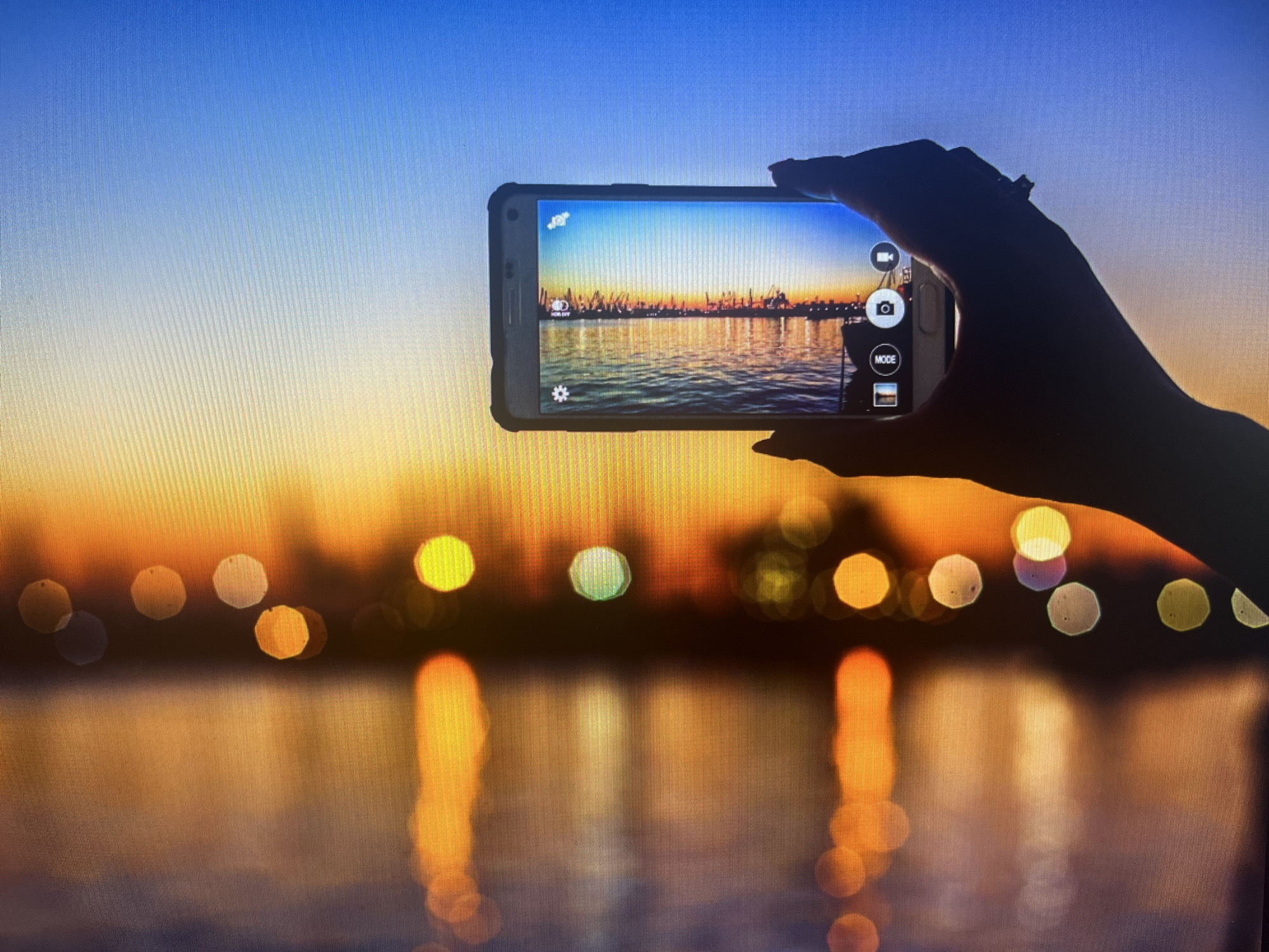 A silhouetted hand holds a smartphone horizontally, capturing an image of a waterfront cityscape at sunset. The sky transitions from deep blue at the top to warm orange and yellow hues near the horizon. Reflected lights from buildings and streetlights create a bokeh effect on the water's surface, appearing as blurred circles of yellow, orange, and white. The smartphone screen displays the scene, with camera interface elements visible, including a video icon, a camera shutter button, and a "MODE" option with a landscape thumbnail. The overall impression is of a serene evening in Varna, Bulgaria, with the city lights illuminating the waterfront as the sun sets.