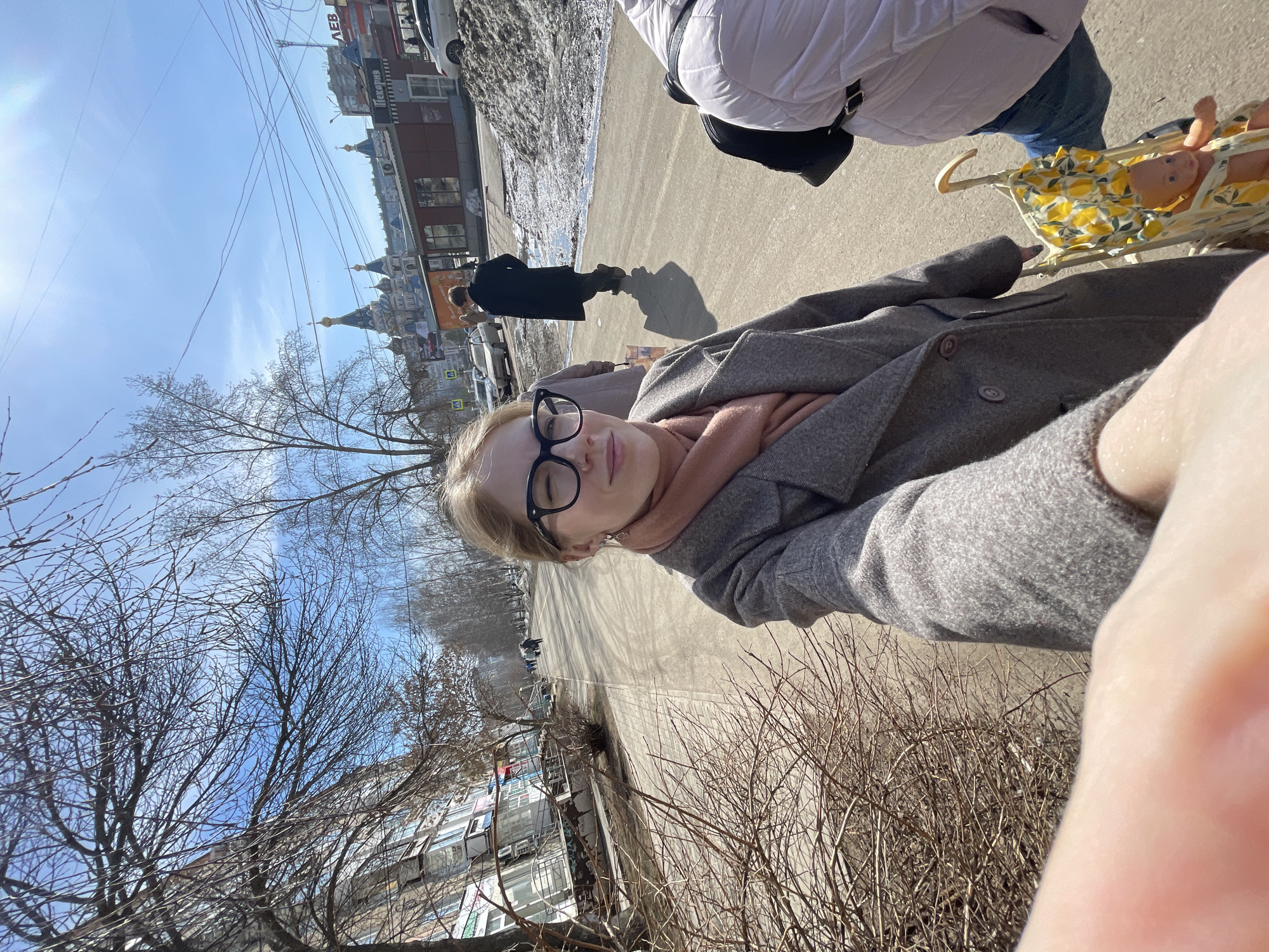A medium-angle shot captures a woman wearing eyeglasses, a gray coat, and a scarf, taking a selfie on a city street. To her right is a person in a white jacket and blue jeans pushing a doll stroller with a doll inside. In the background, a person in a black coat walks along the sidewalk. Buildings are visible in the distance, and the sky is clear blue. The location is Izhevsk, Russia, as evidenced by the cathedral in the background. Bare trees line the street.