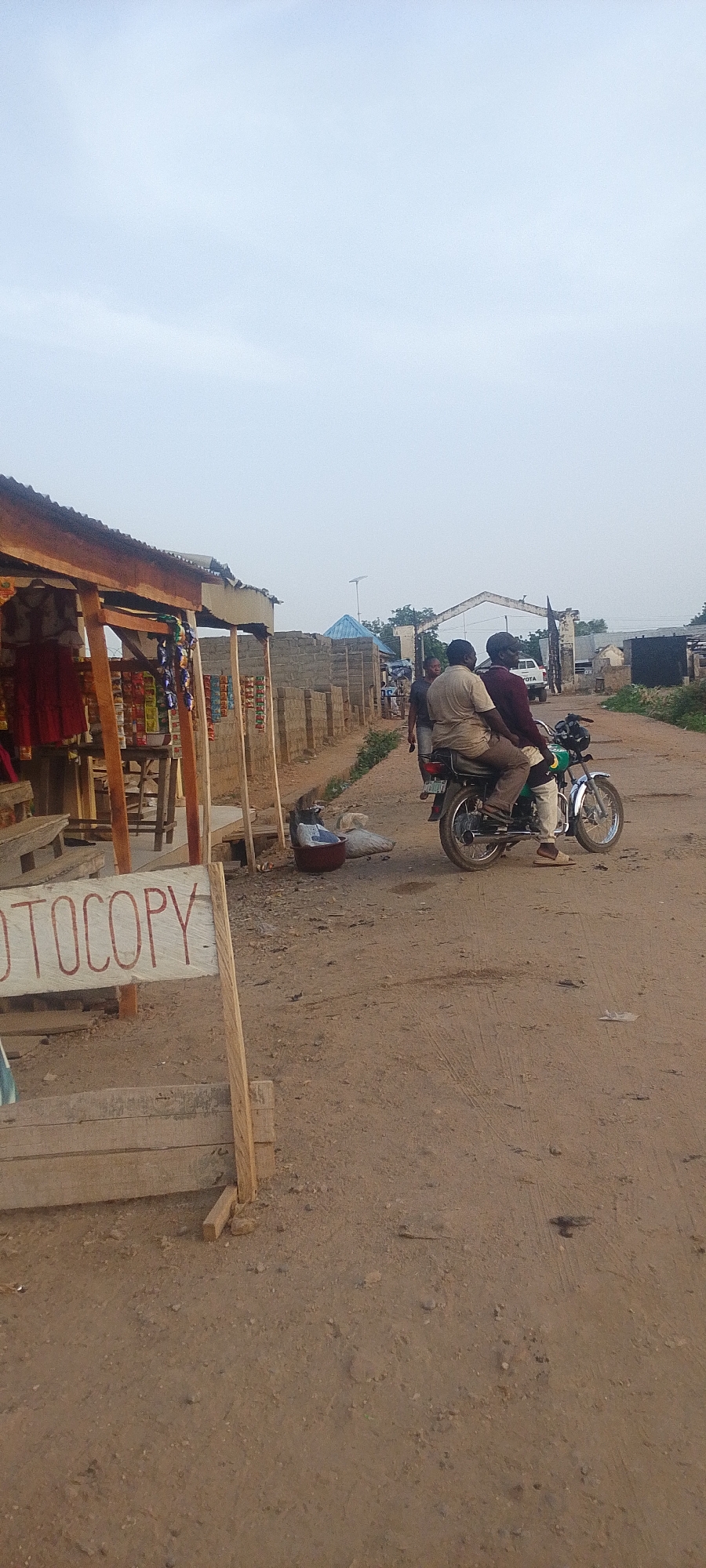 The image depicts a street scene in Adankolo, Nigeria. The unpaved road is dusty. In the foreground, a wooden sign reads "PHOTOCOPY" in front of a small shop with goods displayed. Two men are seated on a green motorcycle in the middle ground. Further down the road, a gate-like structure spans the road, and a white Toyota pickup truck is visible beyond it. Several buildings and structures are scattered along the sides of the road, including some with blue roofs. The sky is overcast. A man is walking behind the motorbike.