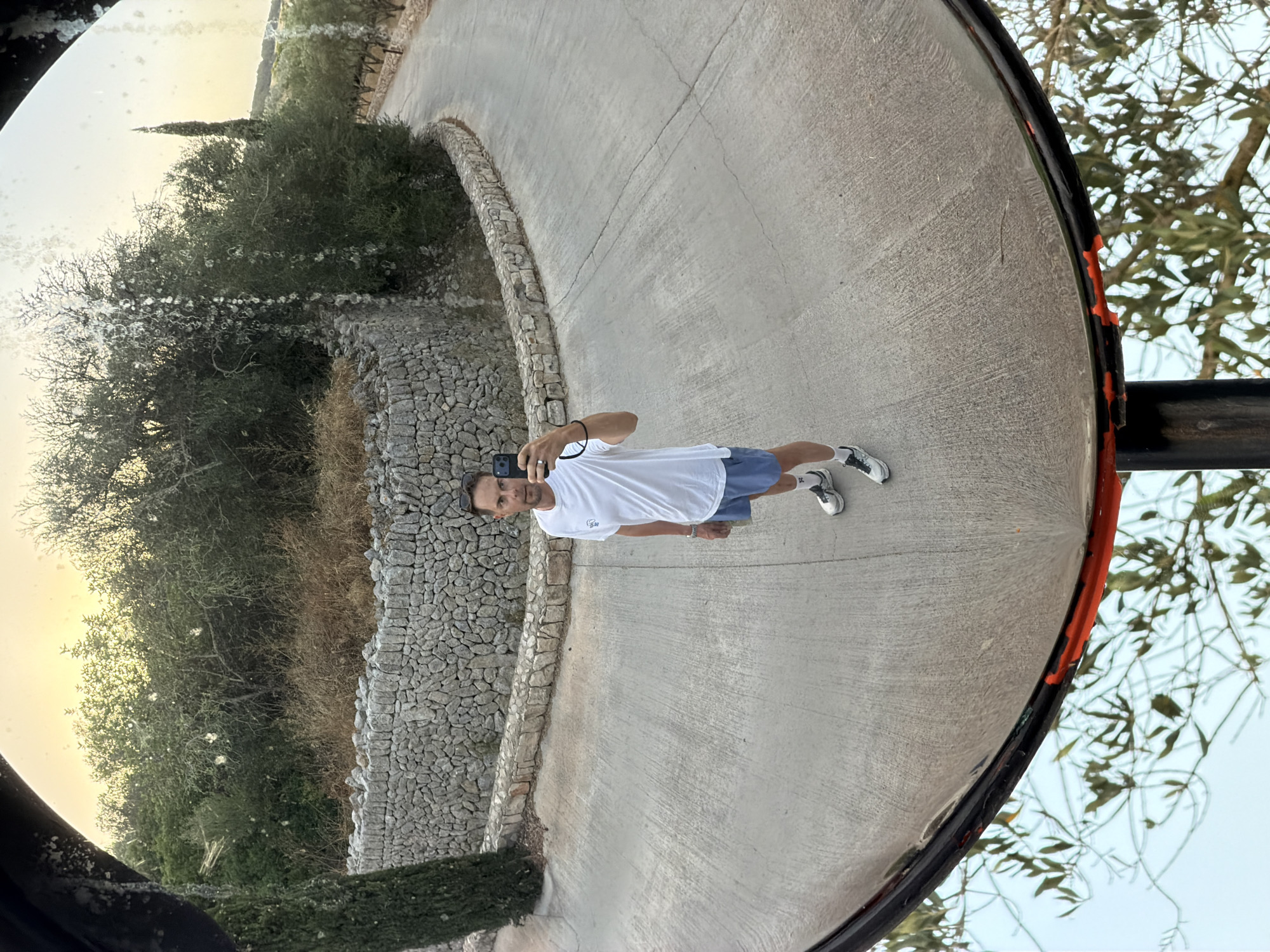 A man is taking a selfie in a convex mirror. He is wearing a white t-shirt, blue shorts, white socks, and athletic shoes. He is standing on a paved road that curves to the right. Behind him, there is a stone wall and dense vegetation. A cypress tree is visible to the left of the wall. The sky is bright, suggesting it is daytime. The scene is set in the city of Montuïri, Spain.