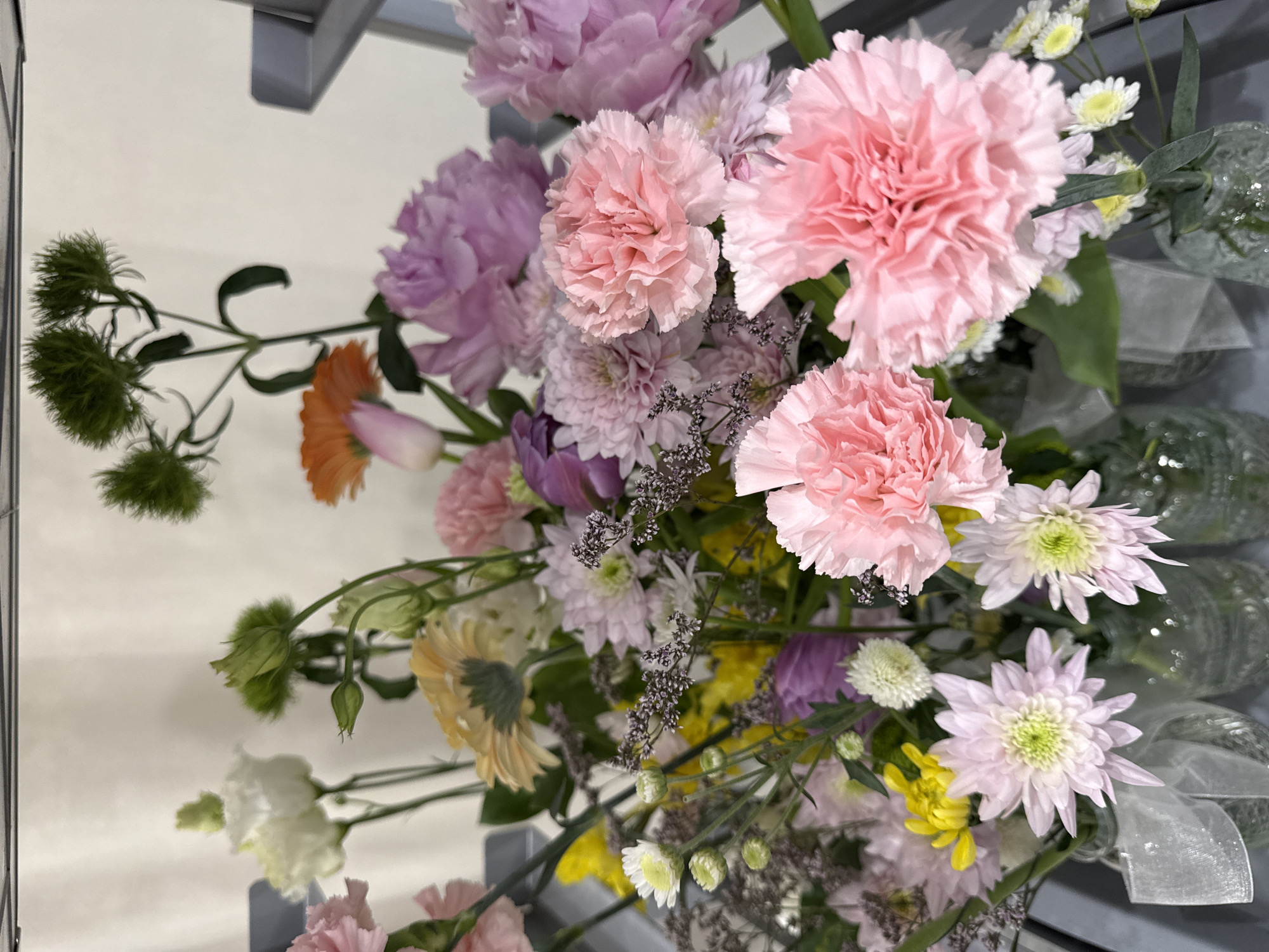 A close-up, angled shot shows a colorful arrangement of flowers including pink carnations, lavender peonies, orange gerbera daisies, and other blossoms in pastel shades. The flowers are likely displayed in a floral shop, possibly in Saint Petersburg, Russia, as the setting suggests an upscale, commercially curated aesthetic with meticulously arranged blooms. Parts of a reflective metal display structure are visible. The overall composition conveys a sense of abundance and visual appeal, typical of floral arrangements intended for purchase.