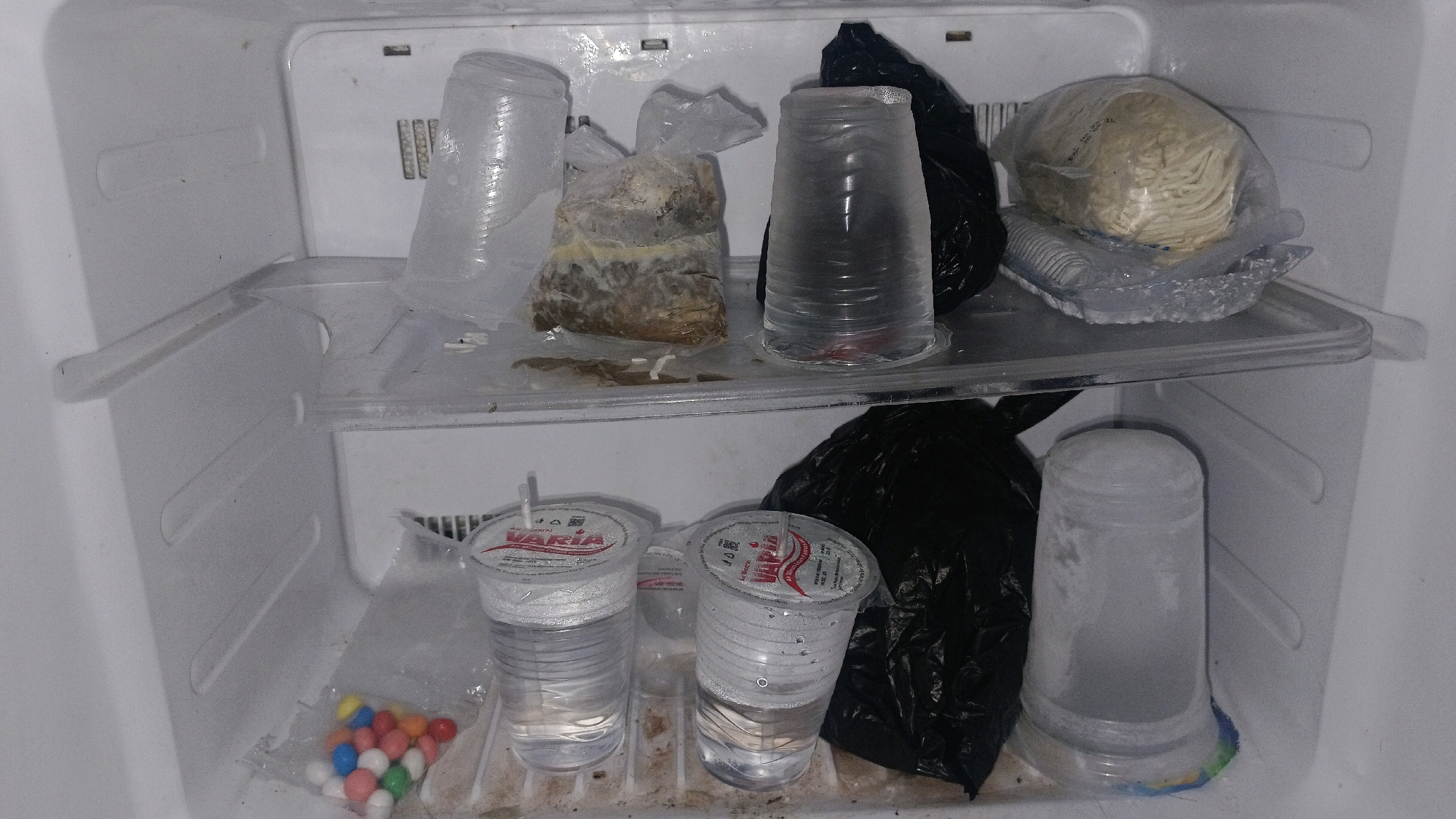 The image depicts the interior of a refrigerator, likely located in a residential kitchen in Central Jakarta, Indonesia. The refrigerator is organized into two shelves. The top shelf contains several items: a clear plastic cup filled with a clear liquid, a plastic bag containing what appears to be food with a yellowish-brown substance, another clear plastic cup with a clear liquid, a black plastic bag, and a plastic bag containing uncooked noodles. The bottom shelf holds two clear plastic cups with straws and clear liquid, a black plastic bag, and a clear plastic cup containing a white, frosty substance that appears to be frozen or ice. In the lower left corner, a small plastic bag filled with colorful, spherical candies is visible. The refrigerator interior appears slightly messy, with some dust or residue visible on the shelves.