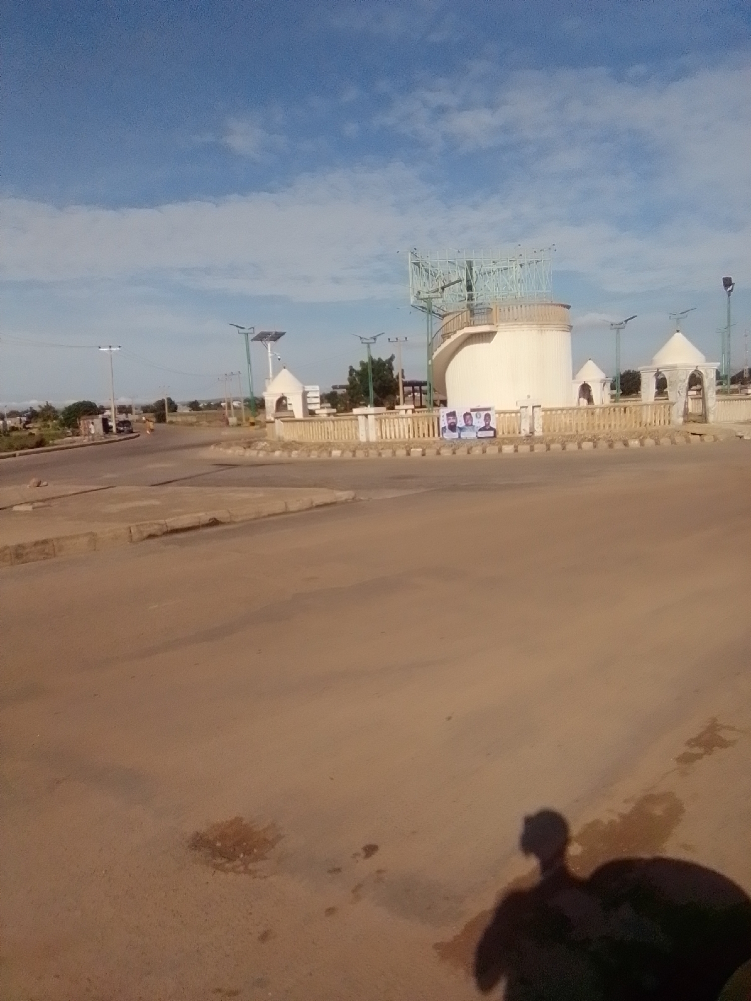 In Kwadon, Nigeria, a circular intersection features a monument at its center. The monument is a multi-tiered white structure with arched openings and a railing. A spiral staircase is visible on the left side of the monument, leading up to a large, open metal framework. In front of the monument, a beige fence separates it from the road. A banner displaying three portraits is hung on the fence. The surrounding environment consists of a paved road, sidewalks, streetlights with solar panels, and scattered trees. The sky above is blue with scattered white clouds. A person's shadow is cast on the road in the foreground, suggesting the presence of an individual out of frame.