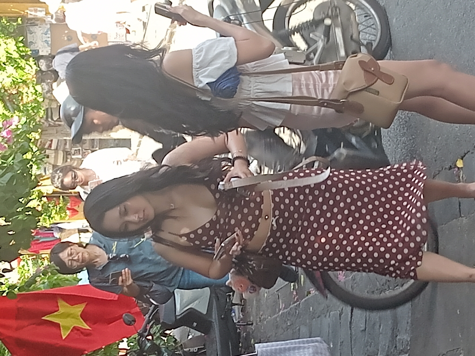 The image, taken in Hoi An, Vietnam, captures a street scene with several people and objects. In the foreground, a woman with long dark hair wears a maroon and white polka dot dress. She is looking down, possibly at something in her hands. To her left, a Vietnamese flag with a yellow star on a red background is visible. Behind her, another woman with long dark hair and bangs is wearing a white dress with blue accents and a small tan purse with a brown strap. She is holding a smartphone. A motorcycle is also present in the background, partially obscured. Further back, another individual, possibly an older woman with glasses, is seen in a blue shirt, also holding a phone. The scene appears to be outdoors, with foliage and what looks like a brick wall in the background. The lighting suggests it is daytime. The overall atmosphere seems casual, possibly a market or a street side gathering.