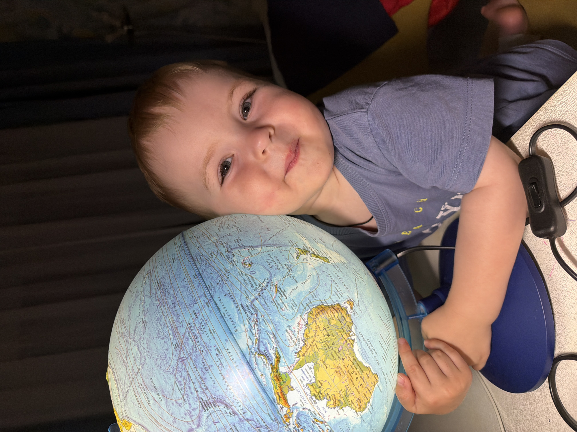 In a dimly lit indoor environment, a young Caucasian child with light brown hair and rosy cheeks is positioned on the right side of the frame, looking towards the viewer with a gentle smile. The child is wearing a light blue t-shirt. The child's left arm is wrapped around a globe that is illuminated from within and rests on a blue stand. The globe displays various landmasses and bodies of water, with text in Russian indicating geographical locations. The child's fingers are touching the surface of the globe. In the foreground, a black cord with an on/off switch is visible on a white surface, likely a desk or table. The background consists of dark curtains, suggesting the room is likely a home. The image appears to be taken in Votkinsk, Russia, given the presence of Russian text on the globe.