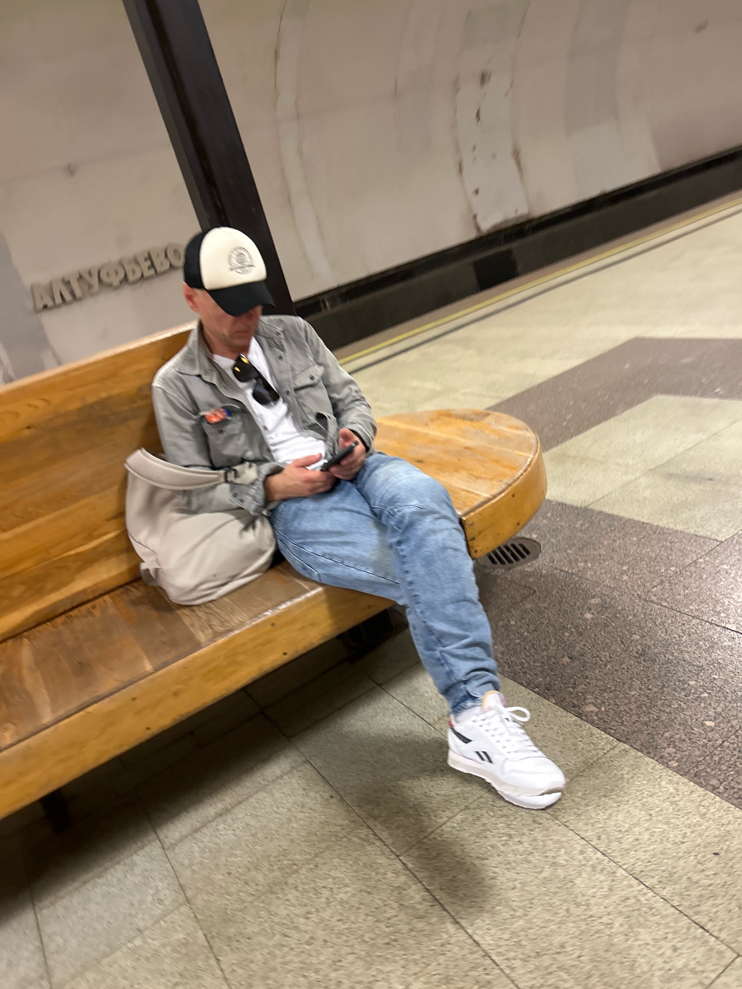 A man is sitting on a wooden bench in a Moscow Metro station, looking at his phone. He is wearing a grey jacket over a white t-shirt, blue jeans, a black and white baseball cap, and white sneakers. A beige bag is placed on the bench next to him. The station features a wooden bench, tiled floor, and a sign with Cyrillic lettering that reads "АЛТУФЬЕВО", indicating the Altufyevo station on the Serpukhovsko-Timiryazevskaya Line. The lighting is dim, characteristic of an underground metro environment.