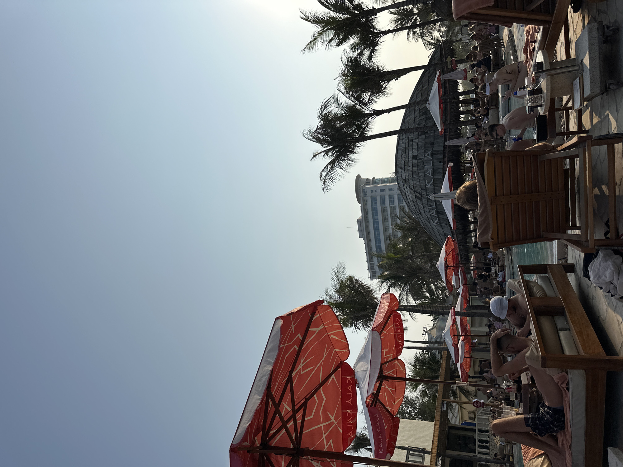 The scene is a poolside area at the Hyatt Regency Đà Nẵng Resort and Spa in Đà Nẵng, Vietnam. Several people are relaxing on lounge chairs next to a pool. Red and white umbrellas provide shade. Palm trees are visible in the background, along with a building that appears to be part of the resort.