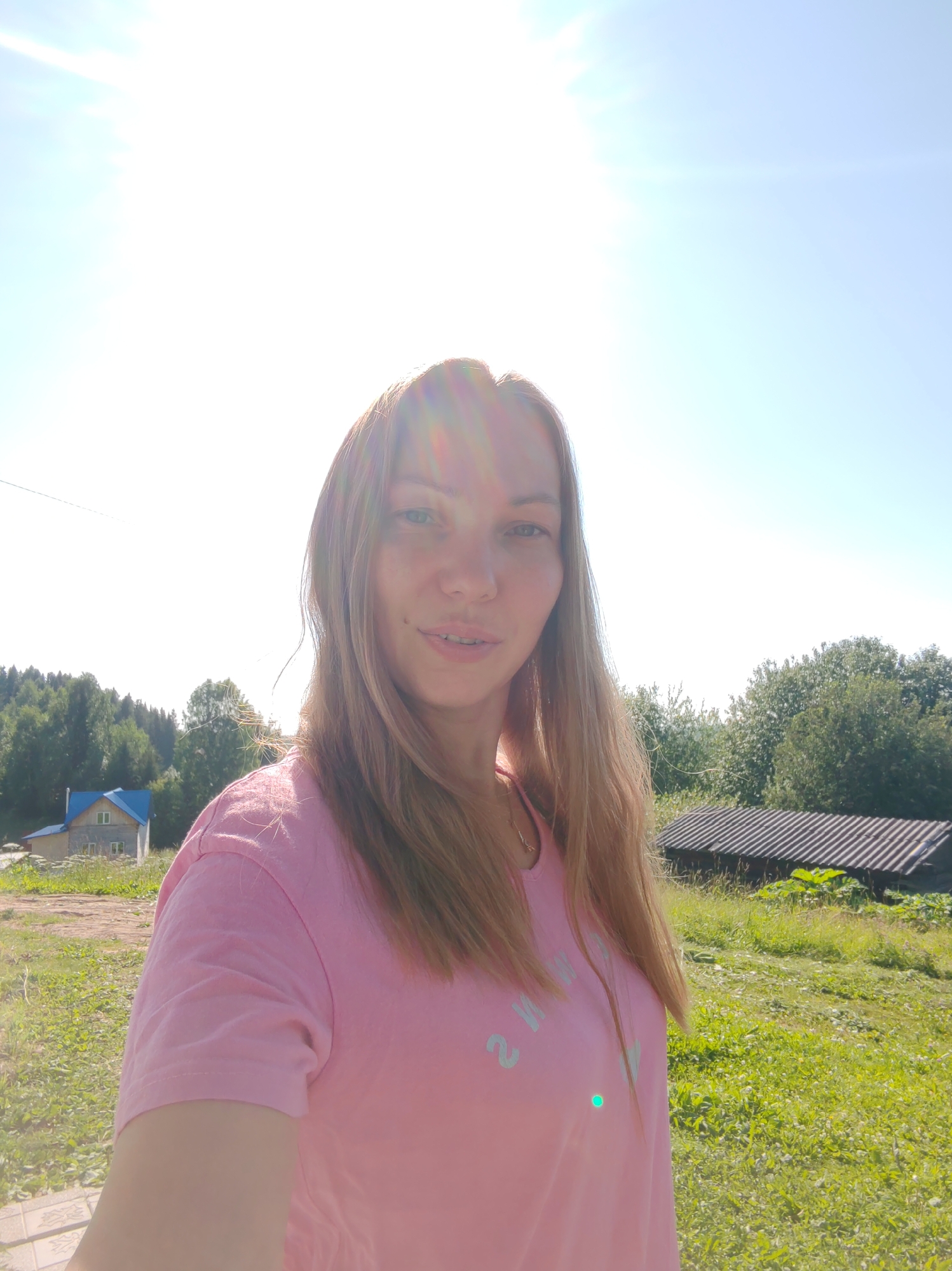 A woman with long blonde hair, wearing a pink t-shirt, stands outdoors in a rural setting on a sunny day. The background features a clear blue sky with a bright sun, a green grassy field, trees, a small house with a blue roof, and a corrugated metal structure. The woman is looking towards the camera.