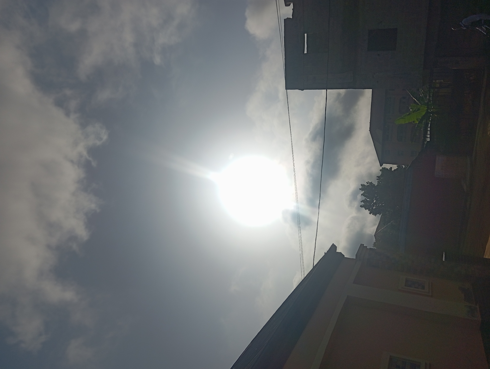 The image shows a low-angle view in Nkpor, Nigeria, looking up at a bright sun partially obscured by clouds. The sky is overcast, with varying shades of gray indicating different cloud densities. Several buildings are visible, including one with a distinctive window. Wires run across the sky, intersecting with the buildings and adding a layer to the urban landscape. A banana tree is present, adding a touch of greenery. The overall impression is of a sunny but overcast day in an urban area.