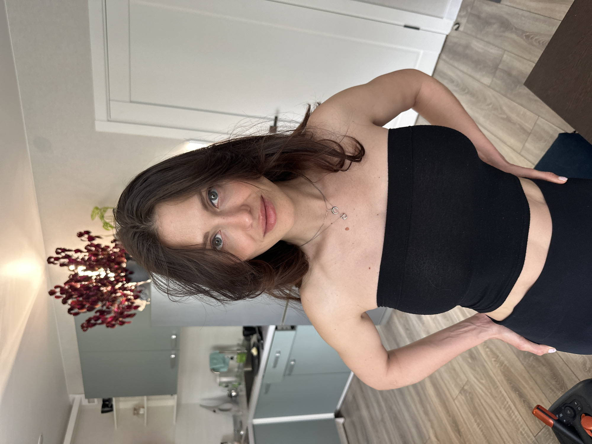 A woman stands indoors, facing the camera at an angle. She wears a black strapless top and matching leggings. Her brown hair falls around her shoulders. She has fair skin and green eyes. Her hands are positioned on her hips. Behind her is a kitchen with light blue cabinets and a white door. There is a floral arrangement in the background. The floor is made of light-colored wood. The location is potentially a residential apartment in Murino, Russia.