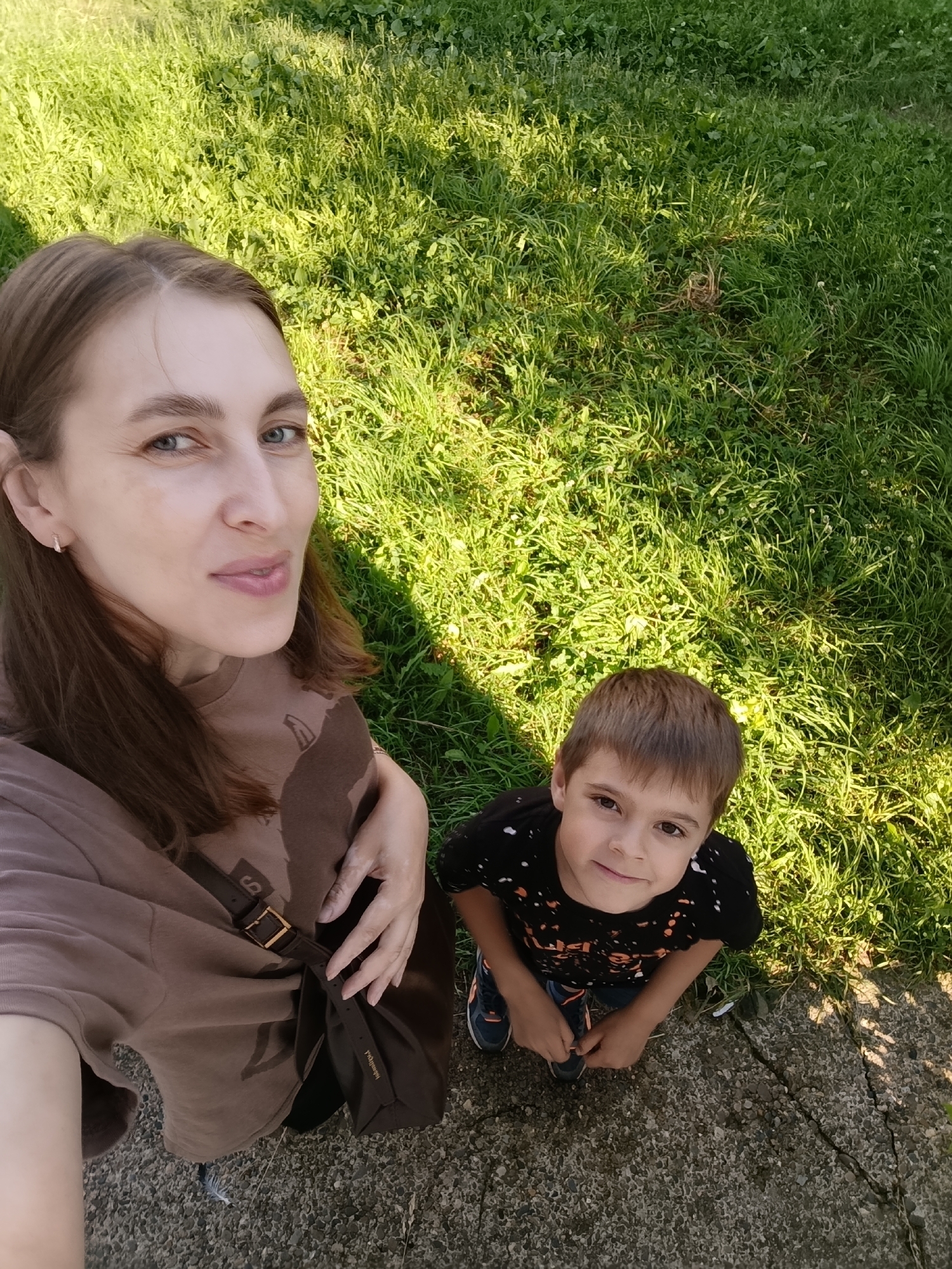 A selfie taken from a high angle shows a woman and a young boy outdoors, with lush green grass in the background and a cracked concrete surface in the foreground. The woman, positioned on the left and in the foreground, has light brown hair, fair skin, and is looking at the camera with a slight smile. She is wearing a brown t-shirt with a dark graphic print and a brown crossbody bag with a gold buckle. Her right hand is resting on her stomach. The boy is on the right side of the image, also looking at the camera with a smile. He has short brown hair and is wearing a black t-shirt with an orange and white splatter pattern and dark sneakers. The lighting suggests it is a sunny day, casting shadows across the grass. The scene appears to be in a park or a yard, consistent with outdoor recreational areas found in cities like Chaikovsky, Russia.