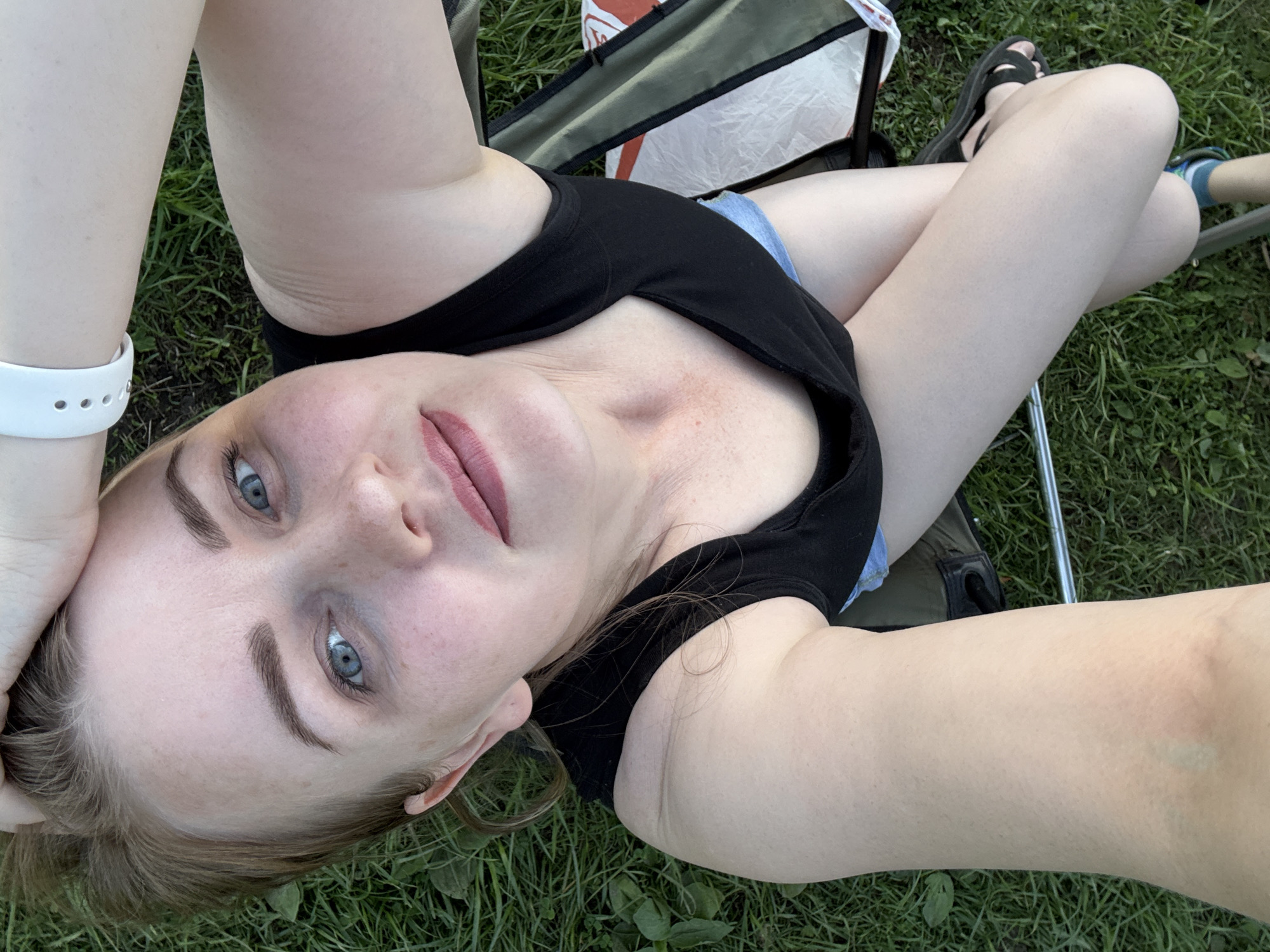 A woman is reclining in a camping chair outdoors, presumably in a park or grassy area. She is wearing a black tank top and blue denim shorts. She is looking at the camera, with her head tilted and one arm raised. The background consists of green grass and the camping chair. There are sandals on her feet. The location is not ascertainable based solely on the provided image.
