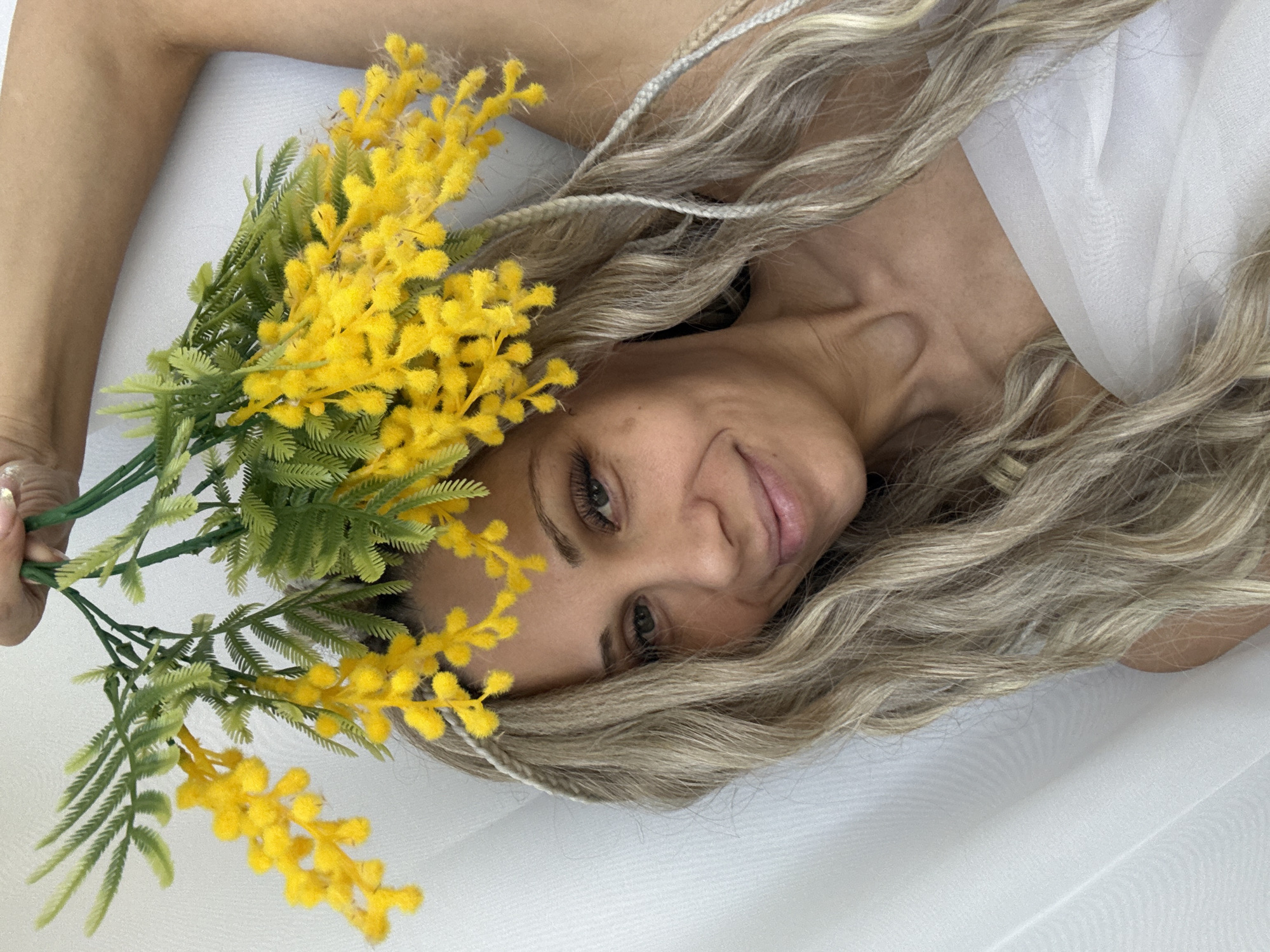 A light-skinned woman with blonde, wavy hair and beaded braids is lying on a white fabric surface, possibly a bed or backdrop. She is smiling and holding a bunch of yellow mimosa flowers with green stems and leaves near her face. The scene appears to be indoors, likely a studio or home setting. The location within the city of Rusanovo, Russia is not determinable from the image.