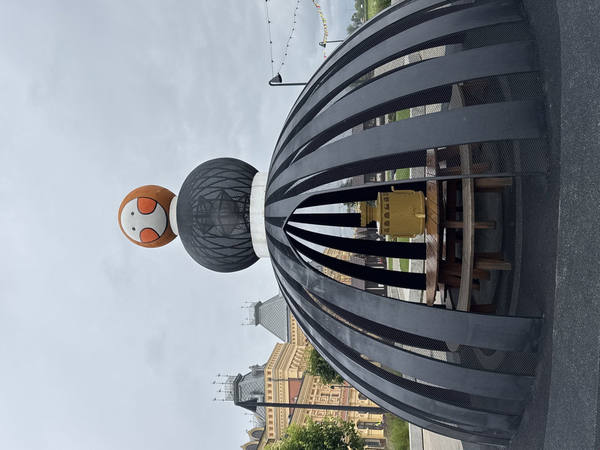 An outdoor scene in Nizhny Novgorod, Russia, features a large, black, dome-like structure with arched ribs and a mesh interior. Within the structure, a wooden picnic table with benches is visible, along with a yellow samovar. Atop the dome, a large, spherical sculpture with a cartoonish face is placed, resembling a stylized character with orange cheeks and black eyes. In the background, ornate yellow buildings with grey roofs and decorative spires suggest an architectural style common in historical Russian cities. The sky is overcast, contributing to the muted lighting of the scene. String lights and small flags are hung in the distance, adding a festive element to the environment. A few trees and green foliage are present, along with paved ground and a darker, textured surface forming the base of the structure. No people are actively engaged in any interactions within the frame.