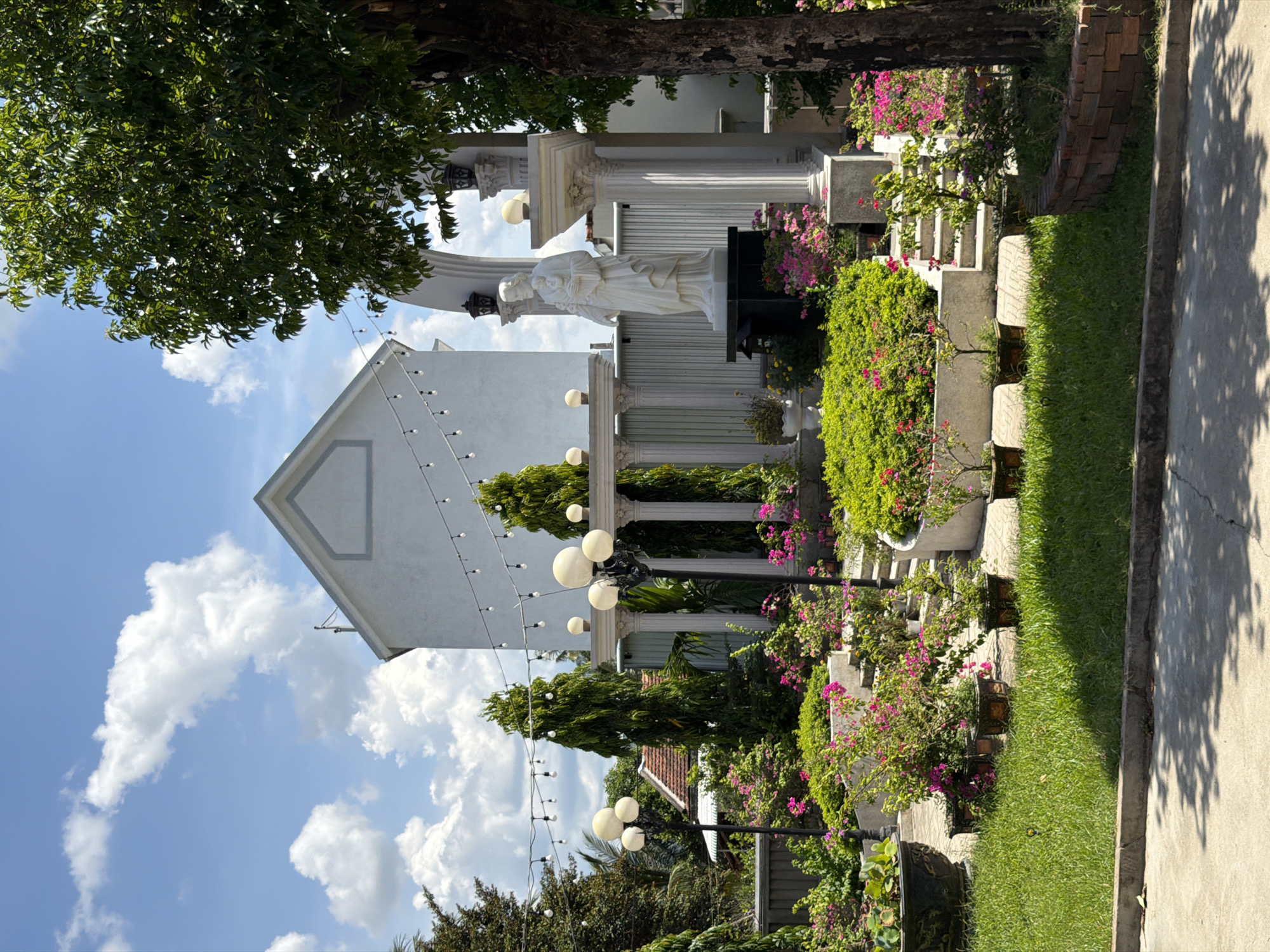 The image depicts a serene outdoor scene in Hội Tín, Vietnam. A white building with a triangular roof is visible in the background, adorned with a string of lights. In front of the building, a white statue of a robed figure holding a child stands on a dark pedestal, framed by white classical columns. The foreground is filled with lush green grass, various potted plants, and vibrant pink flowers. Several tall, slender evergreen trees are strategically placed throughout the garden, adding to the tranquil atmosphere. The sky is a clear blue with scattered white clouds. The overall impression is one of a well-maintained garden or religious sanctuary.