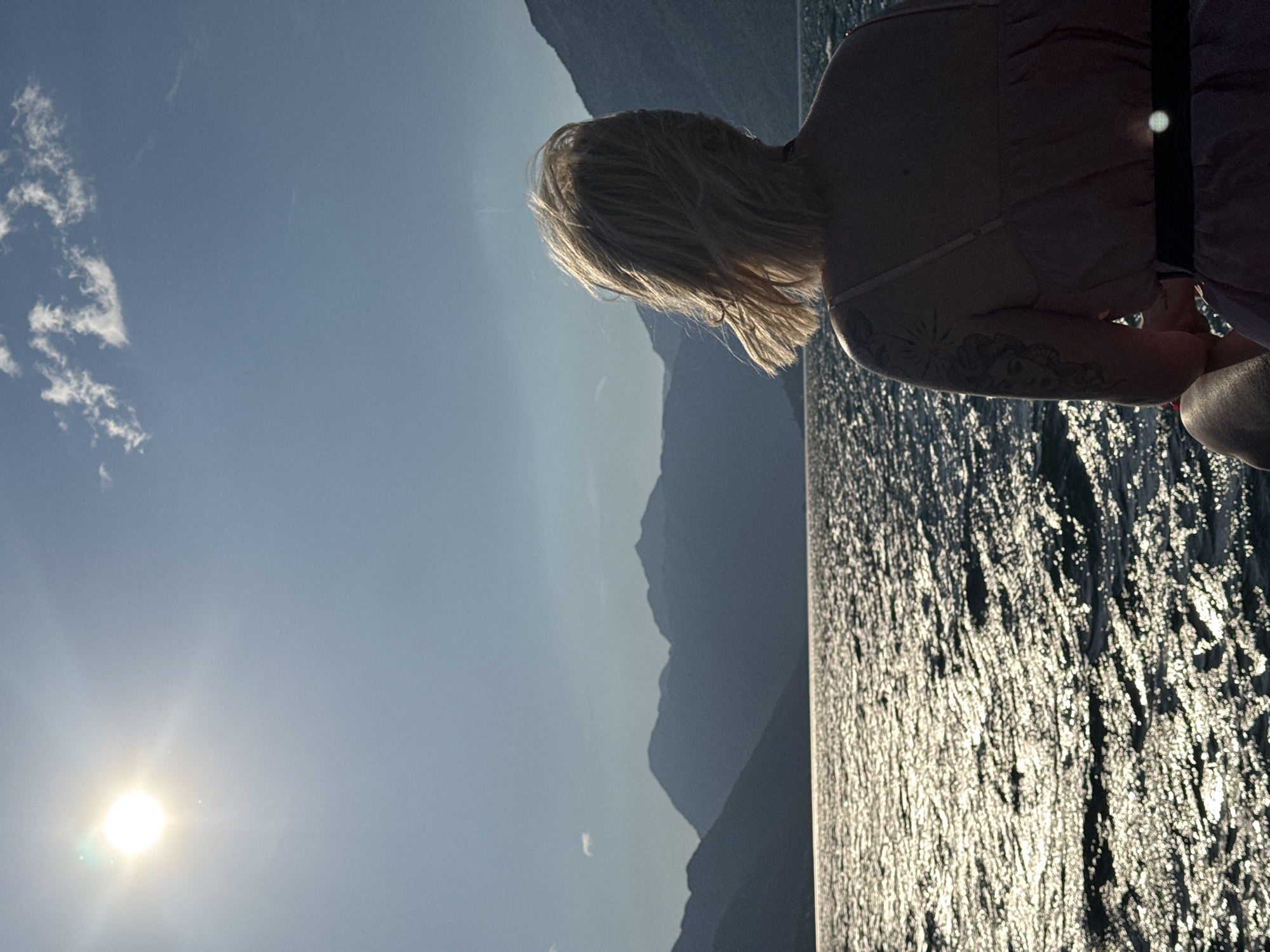 A woman with blonde hair is standing on a boat, looking out over the water. The sun is shining brightly in the sky, creating a sparkling effect on the surface of the water. Mountains are visible in the background. The woman has a tattoo on her arm. This scene is likely in the city of Dobrota, Montenegro, given the location prompt.