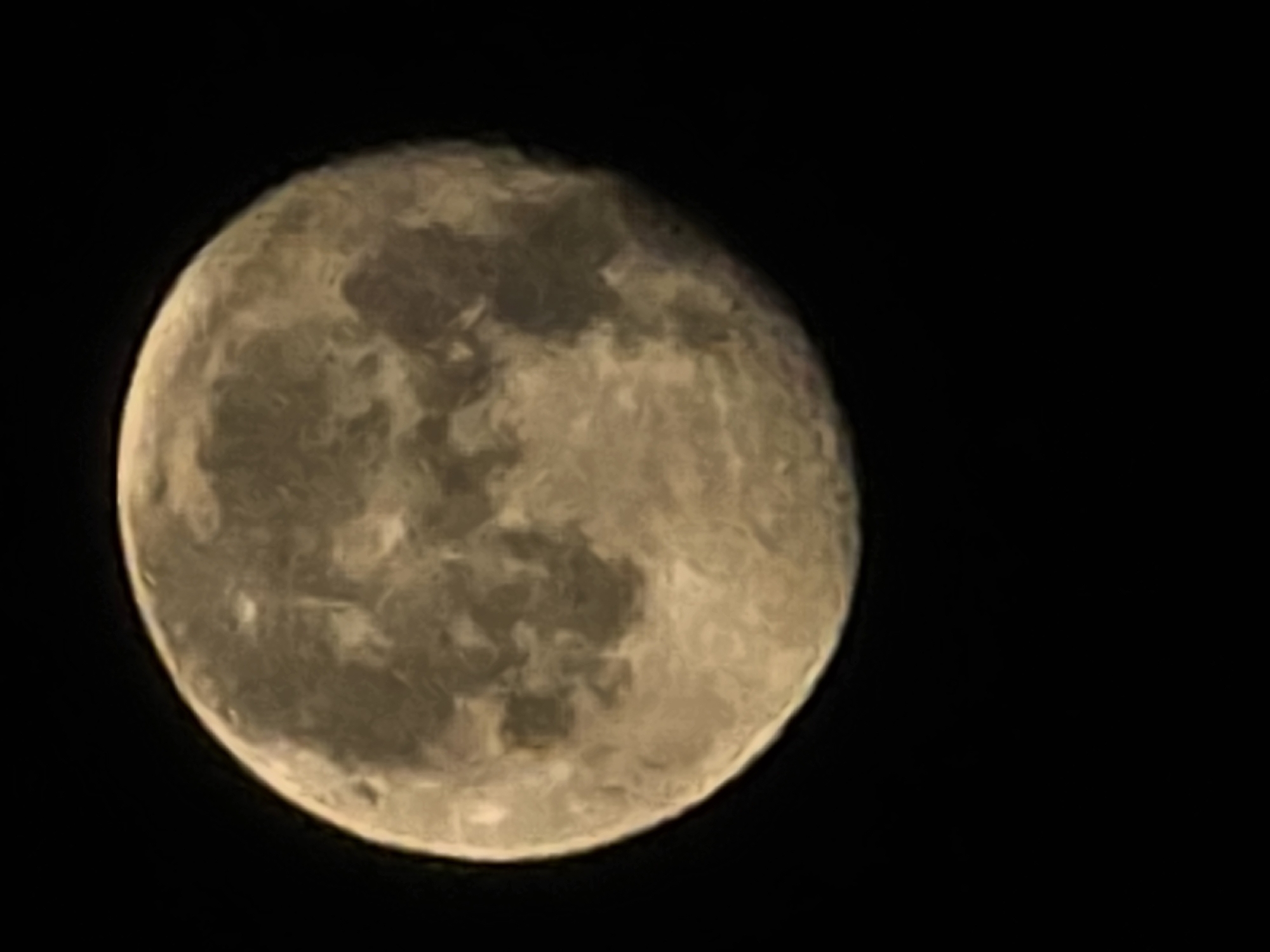 The image shows a close-up view of the moon against a black, starless sky. The moon is illuminated and displays distinct surface features, including craters and darker maria, giving it a textured appearance. The lighting suggests it is either a full moon or very close to it, with a warm, yellowish-brown hue. The image is slightly out of focus, contributing to a soft, ethereal quality. There is no indication of any city or specific location within Yambol, Bulgaria, as the scene is purely celestial.