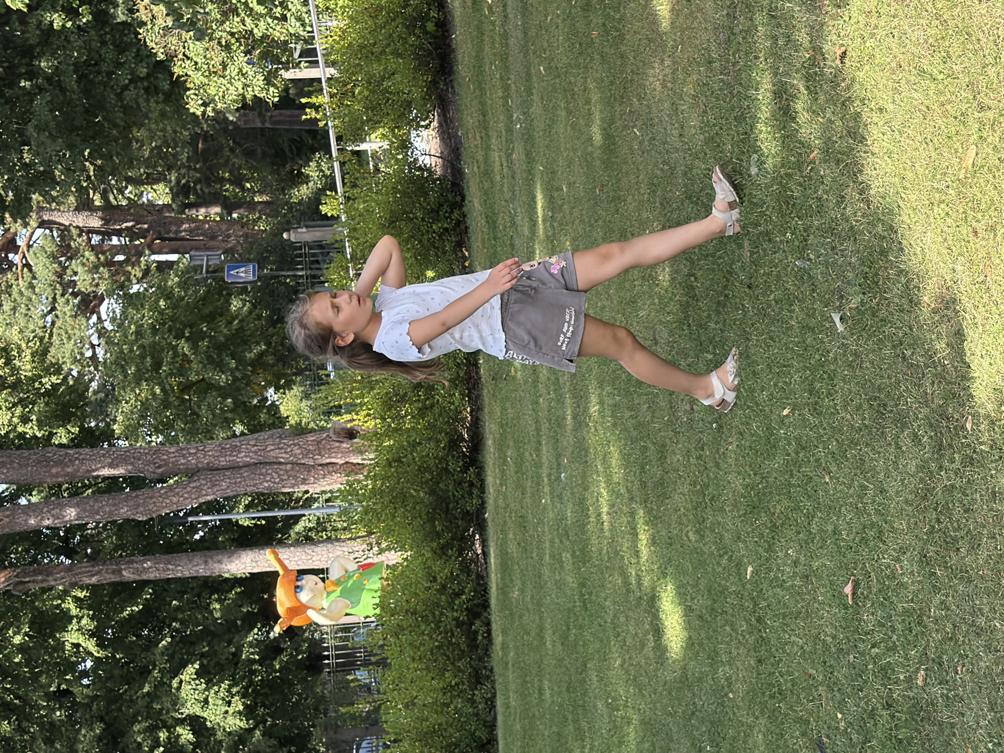 A young girl with blonde hair pulled back in a ponytail is walking on a grassy lawn in a park setting. She is wearing a white t-shirt with a subtle pattern and gray shorts. Her right hand is raised to her head, and she is looking to her left with her eyes closed. She is wearing light-colored sandals. In the background, there are trees, hedges, and a fence. Behind the hedges, a colorful cartoon-like figure with an orange hat and green overalls is visible. The setting appears to be outdoors in a park with mature trees and well-maintained greenery. This scene is located in Ventspils, Latvia.