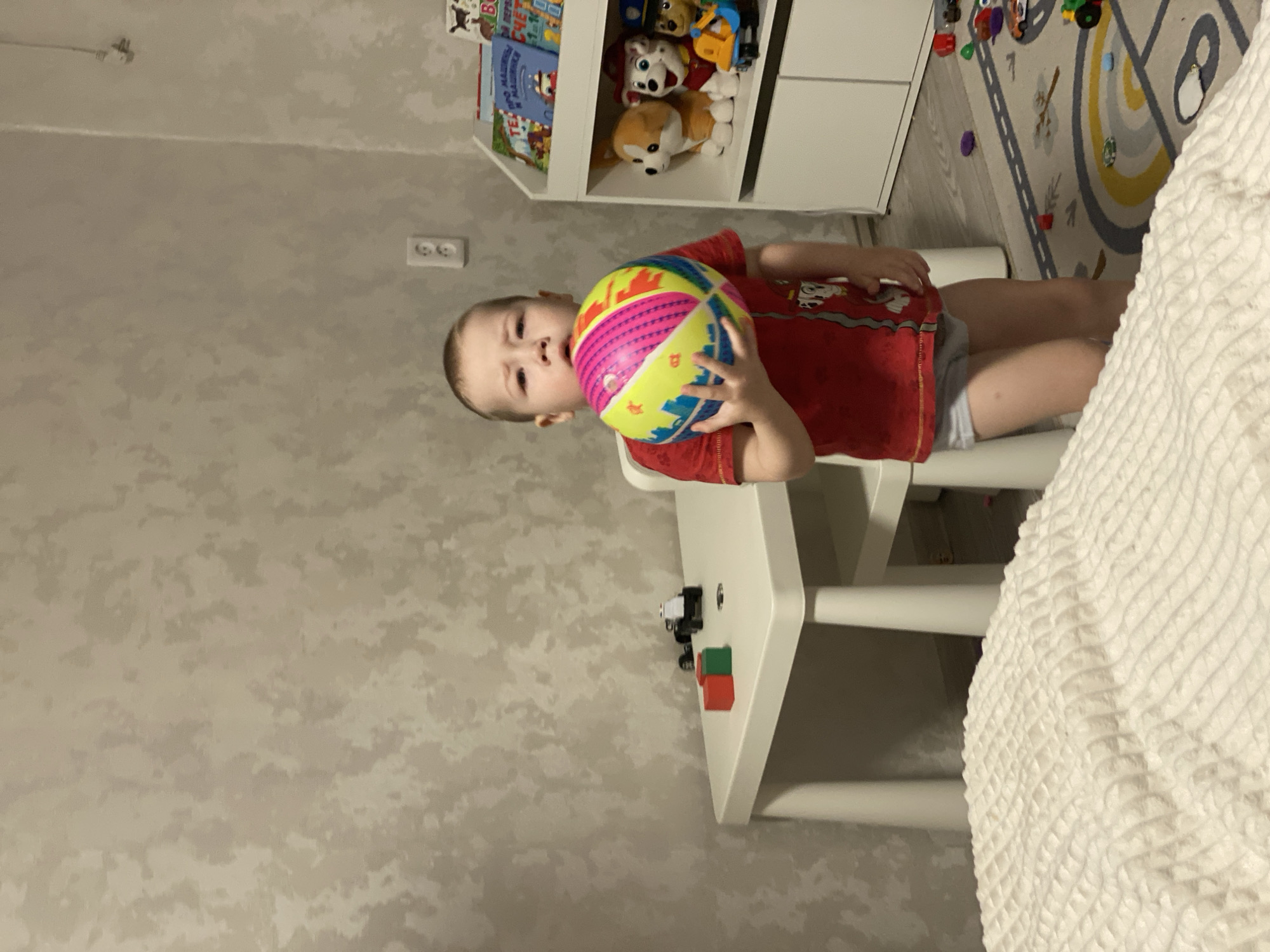 This image depicts a young male child standing in an indoor room, identified as located in Sochi, Russia. The child, with light hair, is positioned centrally, holding a multi-colored spherical ball in both hands, which features pink, yellow, blue, red, and green stripes along with a city skyline graphic. He is dressed in a red short-sleeved t-shirt with a Paw Patrol character graphic on the front and light grey underwear briefs. His expression is a slight frown or pout, with his mouth slightly open, and he is looking directly forward.
To the child's left, a small white rectangular table holds two toy building blocks, one red and one green, along with a black toy vehicle. Directly behind the child, a portion of a small white chair is visible. To the child's right, a white two-tier shelf unit is present. The top tier displays several children's books, some with visible Russian text titles such as "про МАШИНКИ" (about CARS) and "МатЕМАТИКА" (MATHEMATICS). The bottom tier contains multiple plush toys, including a light brown corgi, a white dog wearing a red cowboy hat, and a Paw Patrol character toy in a blue vehicle.
The room's wall features light-toned, mottled wallpaper, and a white electrical outlet with two receptacles is visible on the wall. The lower foreground is occupied by a light-colored, textured blanket or comforter. The floor is covered with a light-colored rug featuring a road map and rainbow arch design, upon which various small toys are scattered.