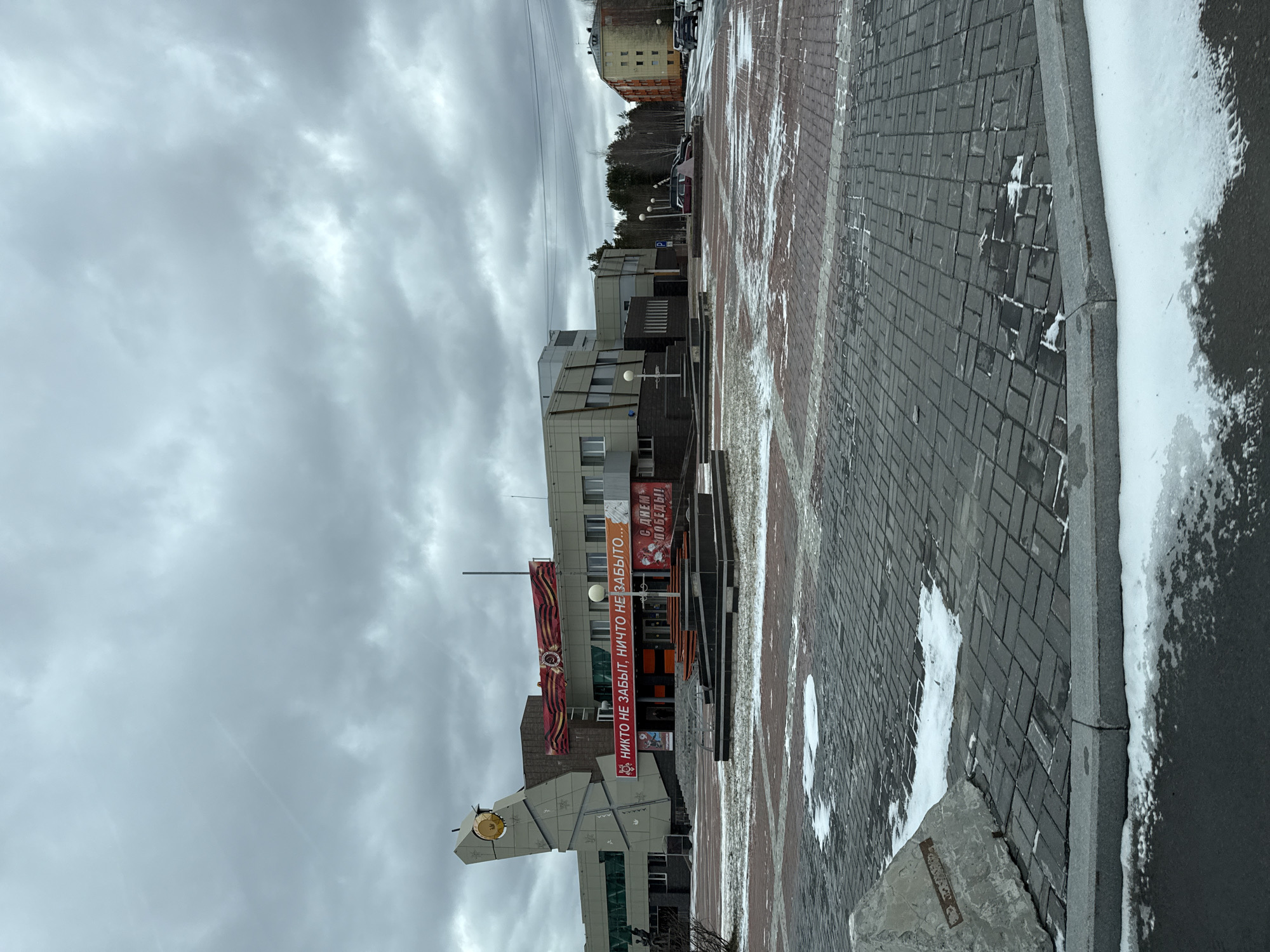 The image depicts an outdoor scene in Beloyarsky, Russia. The sky is overcast with heavy clouds. A modern building with a banner reading "НИКТО НЕ ЗАБЫТ, НИЧТО НЕ ЗАБЫТО..." (meaning "No one is forgotten, nothing is forgotten...") is prominent in the middle ground. The banner appears to be related to Victory Day celebrations. To the left, there's a concrete monument with a clock. The foreground consists of a paved pedestrian area, with some patches of snow and ice visible. In the background, there are more buildings and trees. There are no people visible in the frame.