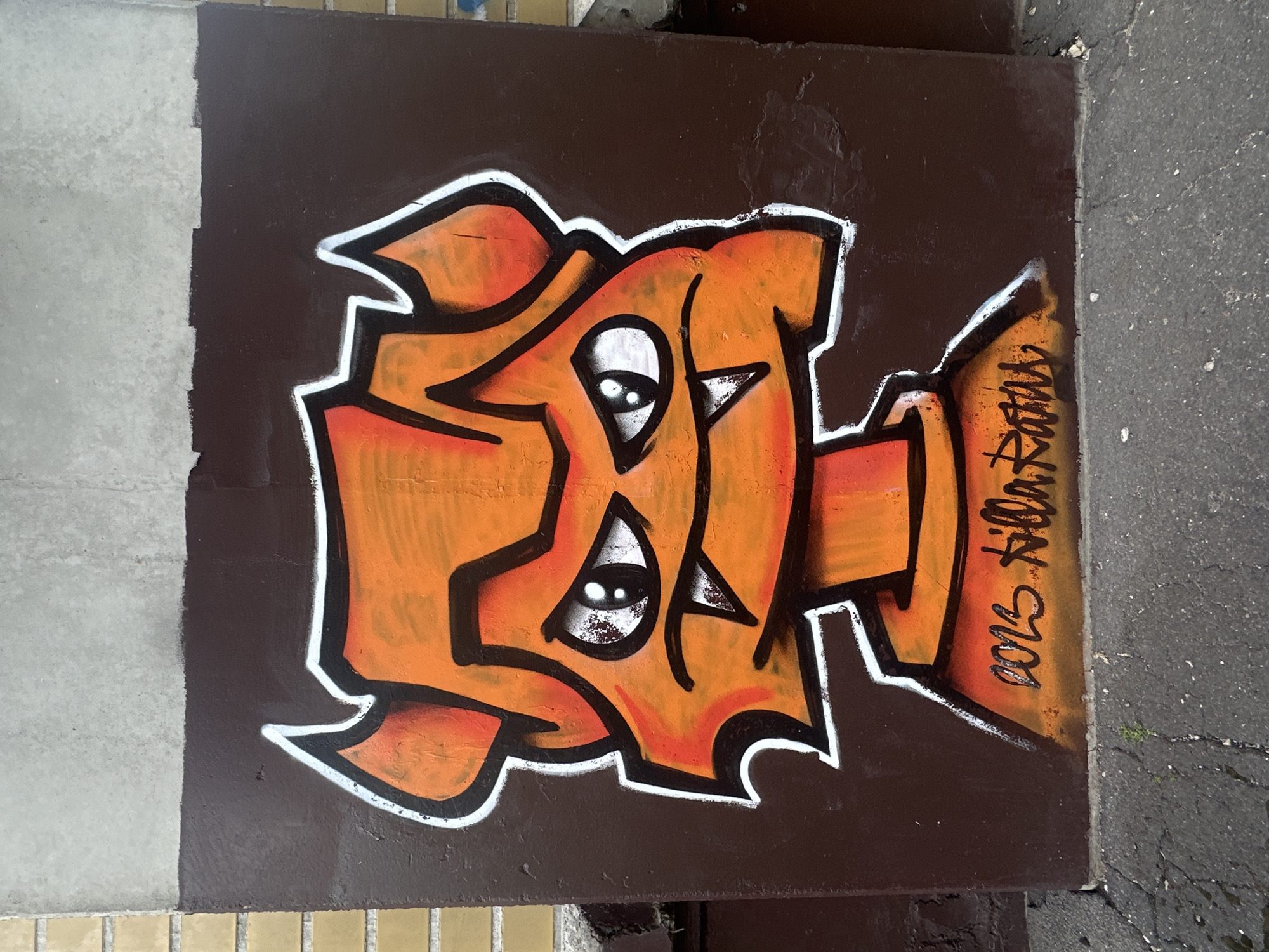 The image depicts a piece of graffiti on a brown concrete structure, possibly a bridge support or wall, in Riga, Latvia. The graffiti is primarily orange with black and white outlining, featuring two stylized eyes within the design. The signature "2013 Killa Ratas" is present on the right side of the graffiti. The structure is bordered by gray concrete on the left and a gray, cracked sidewalk on the right.