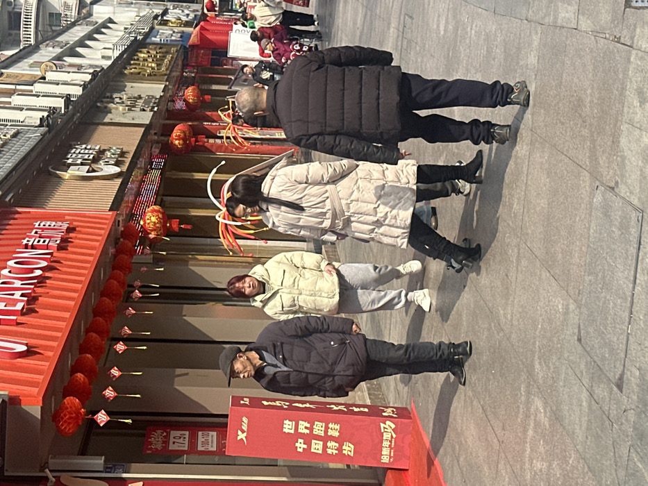 A street scene shows four individuals walking in a line. The first person is an older man in a dark puffer jacket. Behind him is a woman in a long, light-colored coat. She has what appear to be bunny ears or streamers attached to her head. Following her is another woman in a lighter puffer jacket and gray sweatpants. The last person is another older man in a darker jacket and baseball cap. They are walking in front of a building adorned with red signs and decorations.