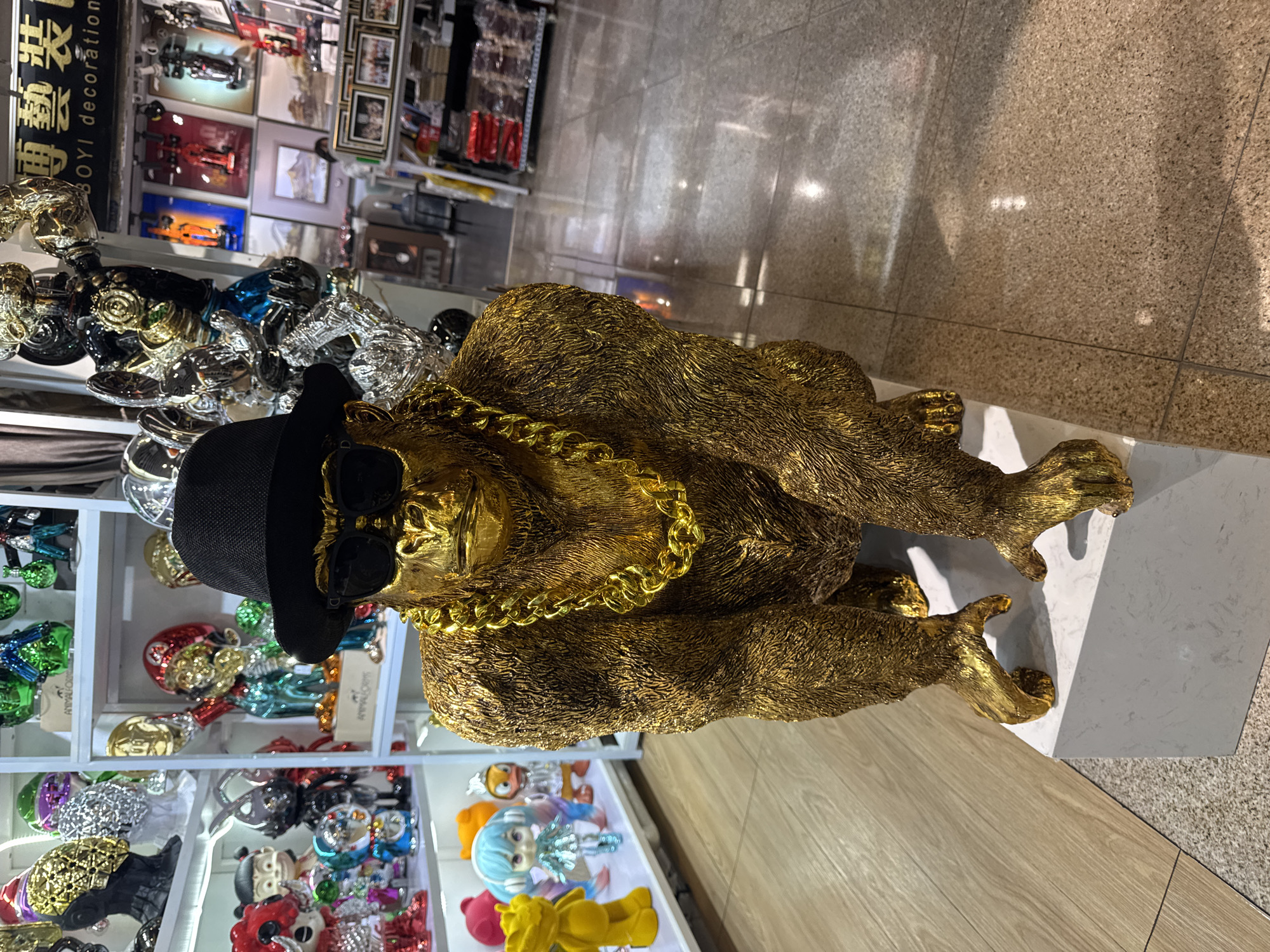 A gold-colored, gorilla statue wearing a black hat and sunglasses and a gold chain is on display at a shop in Yuexiu District, China. The sculpture is on a white marble pedestal. Behind it are shelves stocked with other decorative items, including colorful figurines. The shop has tiled flooring and is brightly lit.
