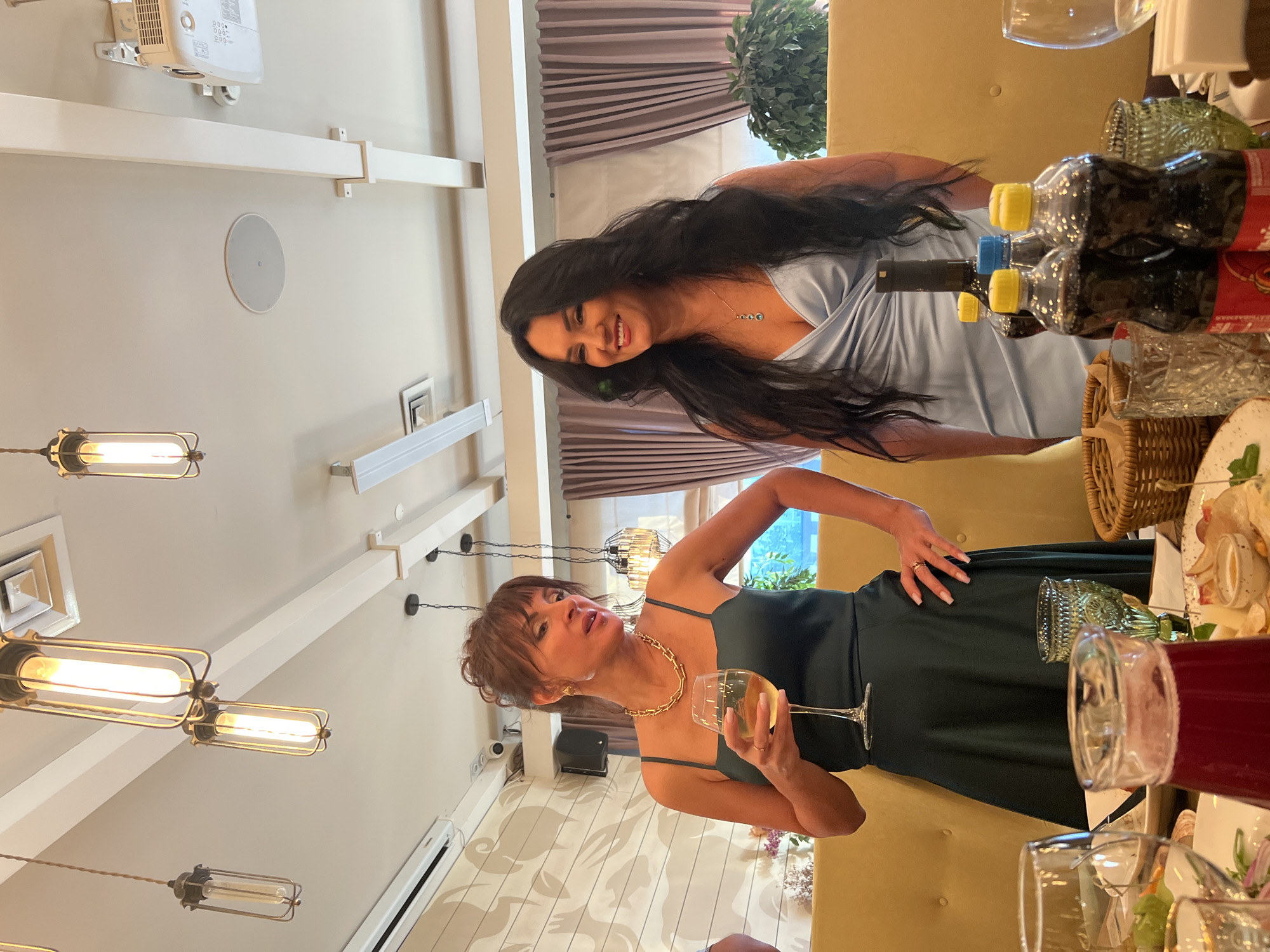 Two women are standing in a restaurant that has a warm, ambient lighting. The woman on the left is wearing a dark green dress and holding a glass of white wine. She has her hair up and is wearing gold jewelry. The woman on the right is wearing a light blue dress and has long, dark hair. She is smiling. In the foreground, there is a table with drinks and food. The environment appears to be a formal gathering or celebration.