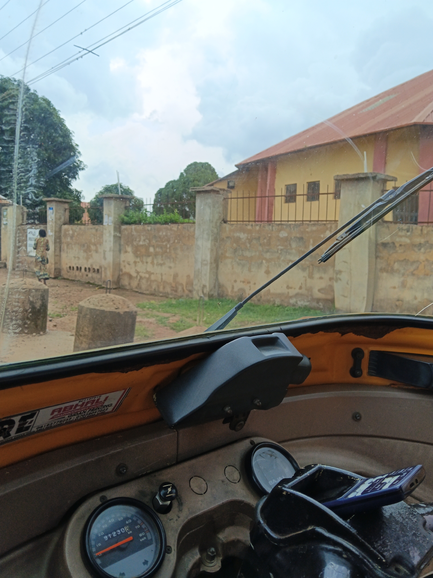 The image shows the interior of a yellow auto-rickshaw, looking out through the windshield. The dashboard features a speedometer with a reading of 91230 km/h, a mobile phone in a holder, and various other controls. Outside the vehicle, a tan-colored wall with openings runs along the street, behind which is a yellow building with a red roof. A person in a patterned dress is visible walking on the street. The sky is overcast with clouds. The location is described as Unknown, Unknown.