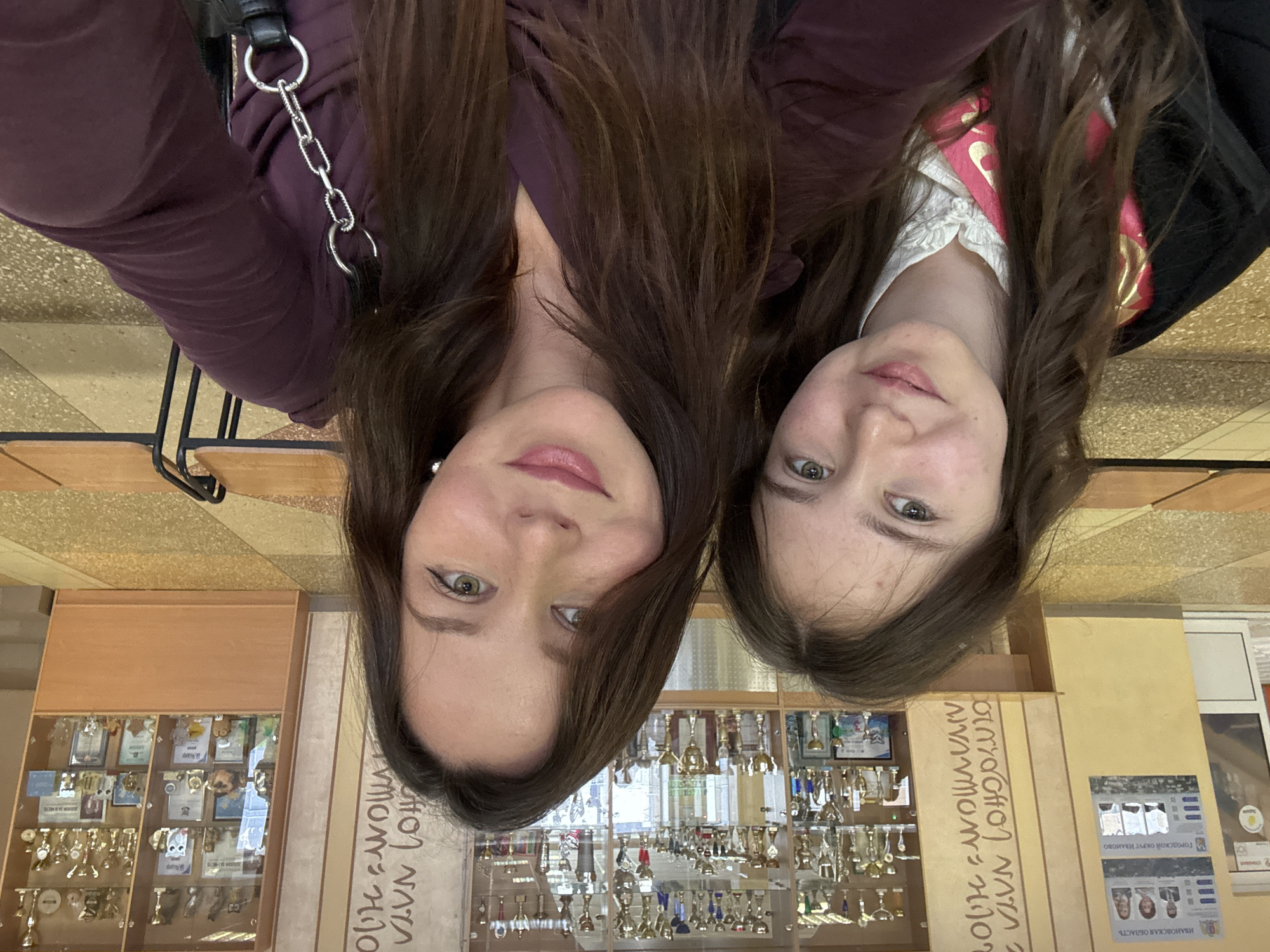 Two women are posing for a selfie in front of a display case filled with trophies, medals, and certificates. The display case has inscriptions in Russian, including "ИВАНОВСКАЯ ОБЛАСТЬ" (Ivanovo Oblast), "ГОРОДСКОЙ ОКРУГ ИВАНОВО" (City District Ivanovo), and "ОТРАСЛЕВОМУ МОЛОДЕЖНОМУ ХОЛДИНГУ NATA" (Sectoral Youth Holding NATA). The woman on the left is wearing a burgundy top with a chain detail, while the woman on the right appears to be wearing a white blouse with ruffles and a red and gold sash. Both women have long brown hair and are looking at the camera. The background features neutral-colored walls and furniture, suggesting an indoor location within a building.