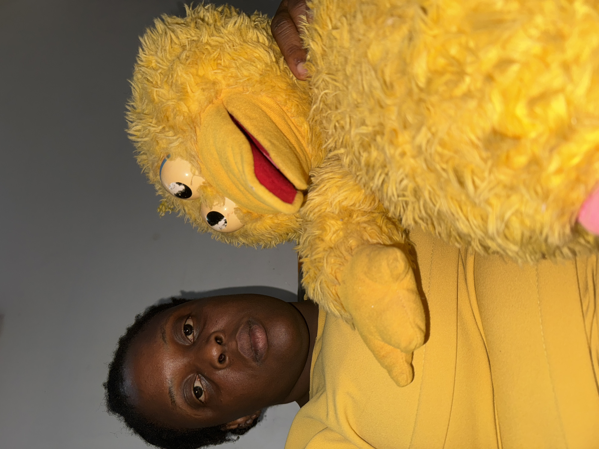 A photograph shows a dark-skinned individual holding a large yellow plush toy with multiple eyes and a red-lined mouth, positioned at a tilted angle in front of a plain, light gray background. The person is wearing a yellow top that matches the color of the toy. The image appears to be taken indoors with artificial lighting. The exact location is Unknown, Unknown.