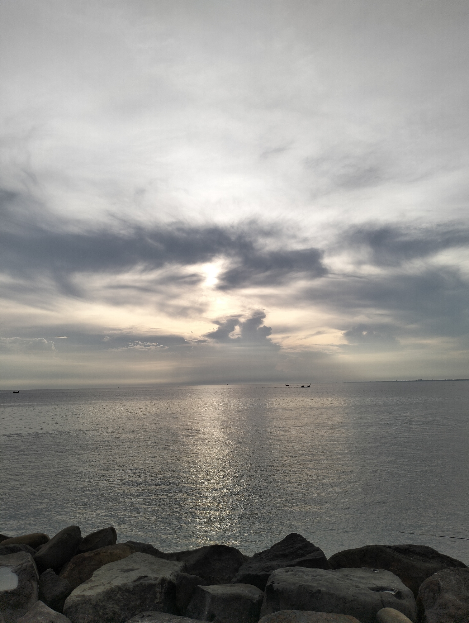 The image shows a seascape in Lhokseumawe, Indonesia. Above a rocky shore in the foreground, the sea reflects a path of sunlight through heavy cloud cover. Three small boats are visible on the horizon. The scene is overcast, with the sun partially obscured by the clouds.