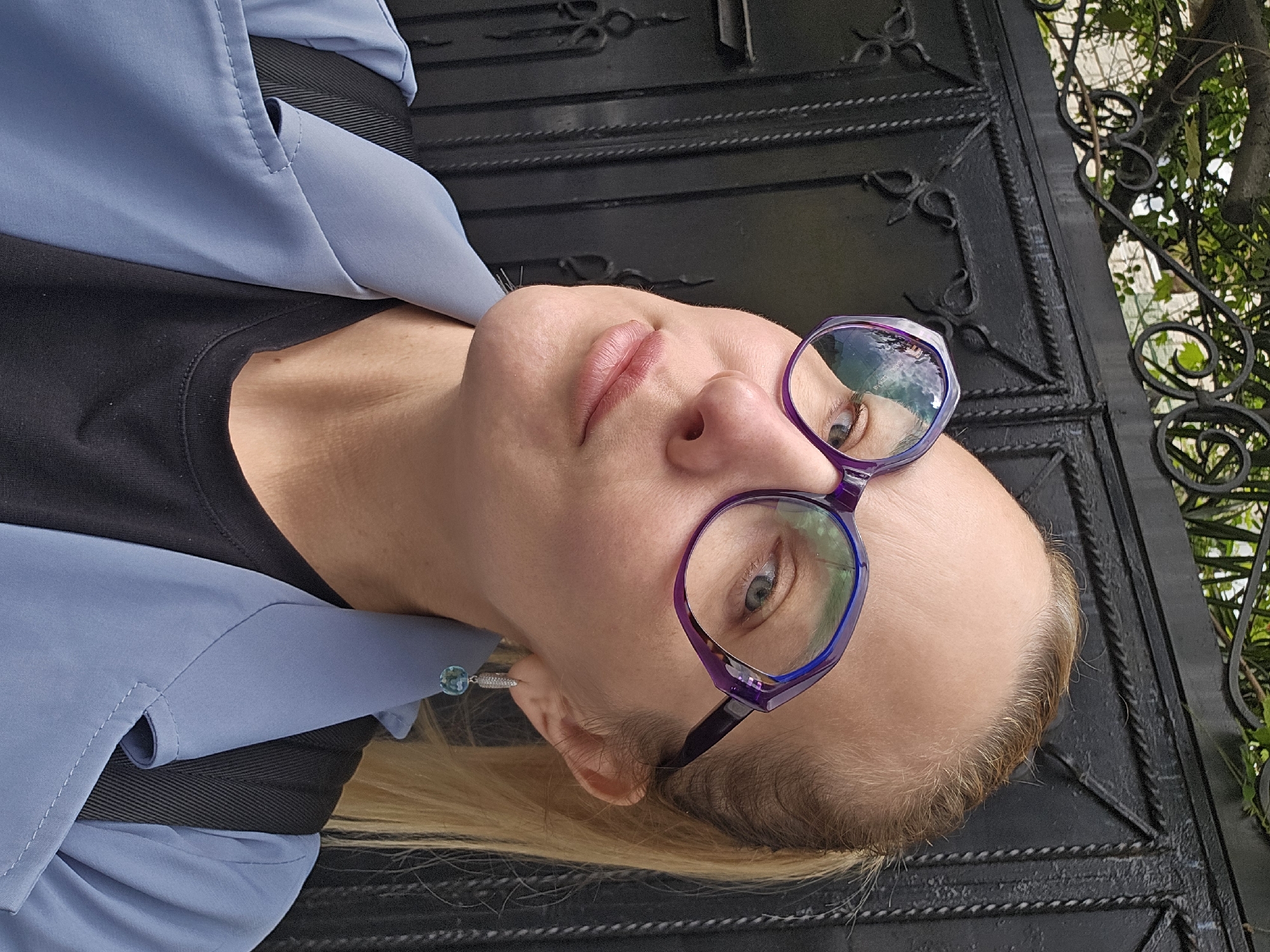 A fair-skinned woman with blonde hair pulled back and wearing purple-framed glasses is looking upwards at the camera. She is wearing a black shirt under a light blue jacket, with a black strap visible over her shoulder. The background consists of a dark, ornate metal gate and green foliage. The location is Sochi, Russia.