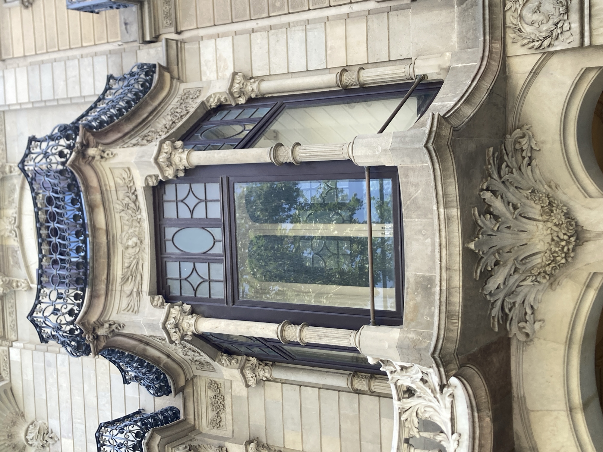 This is a low-angle outdoor photograph of a bay window on a building in Barcelona, Spain. The building's facade is made of light-colored stone with ornate architectural details. The bay window is large and features dark-colored window frames with a geometric pattern in the glass. There are balconies with black wrought-iron railings above and to the sides of the bay window. The building exhibits classical architectural elements, including columns with decorative capitals and sculpted reliefs. The overall impression is one of grandeur and historical architecture.